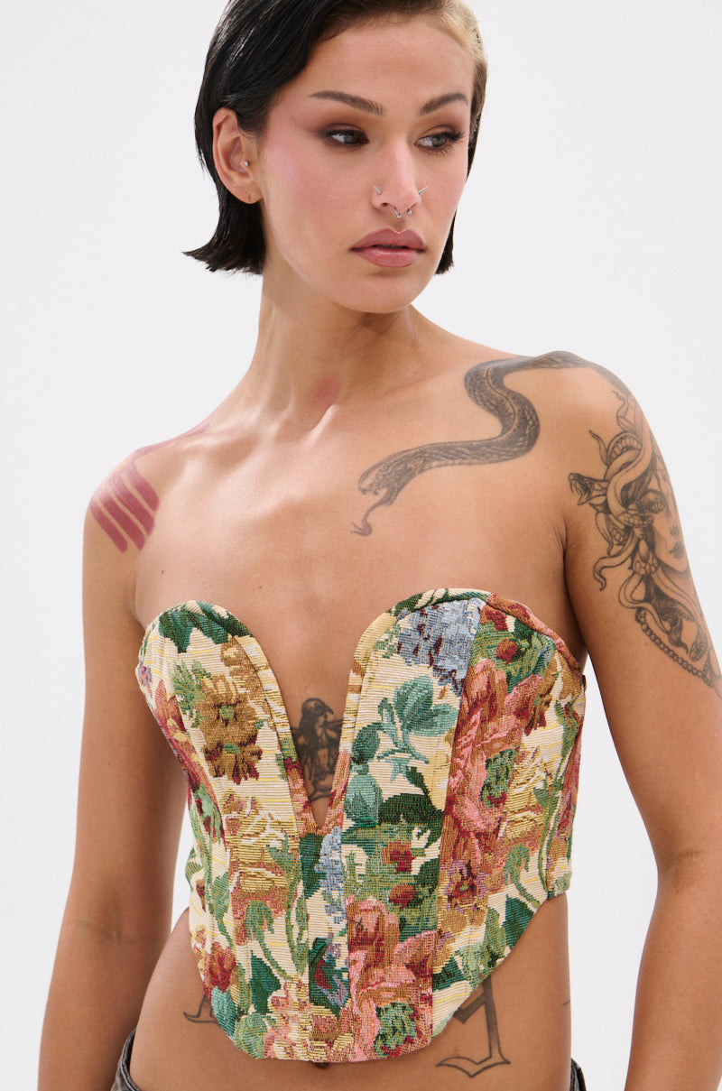 A person with short dark hair and tattoos models the BRIGETTE DISTRESSED JACQUARD CORSET in multicolored floral tapestry, featuring a strapless sweetheart neckline, while looking to the side against a plain light background.