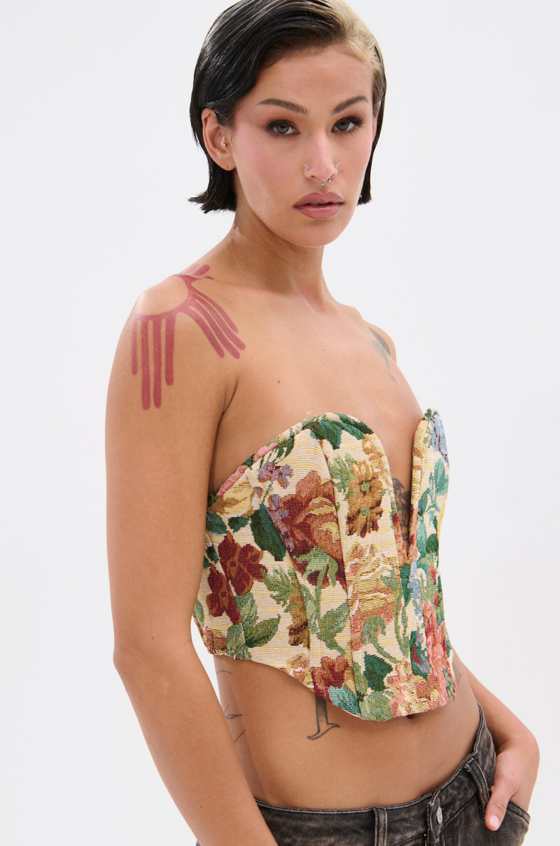 A person with short dark hair and bold makeup wears the BRIGETTE DISTRESSED JACQUARD CORSET and dark jeans. They have a pink abstract shoulder tattoo and a nose ring, standing against a plain white background.
