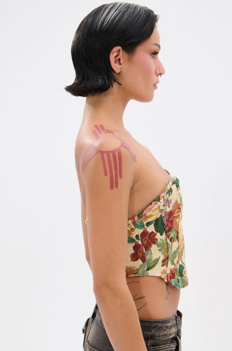 A person with short black hair stands in profile, wearing the BRIGETTE DISTRESSED JACQUARD CORSET and dark pants. They display bold pink abstract body paint on their shoulder and arm, along with visible tattoos on their side.