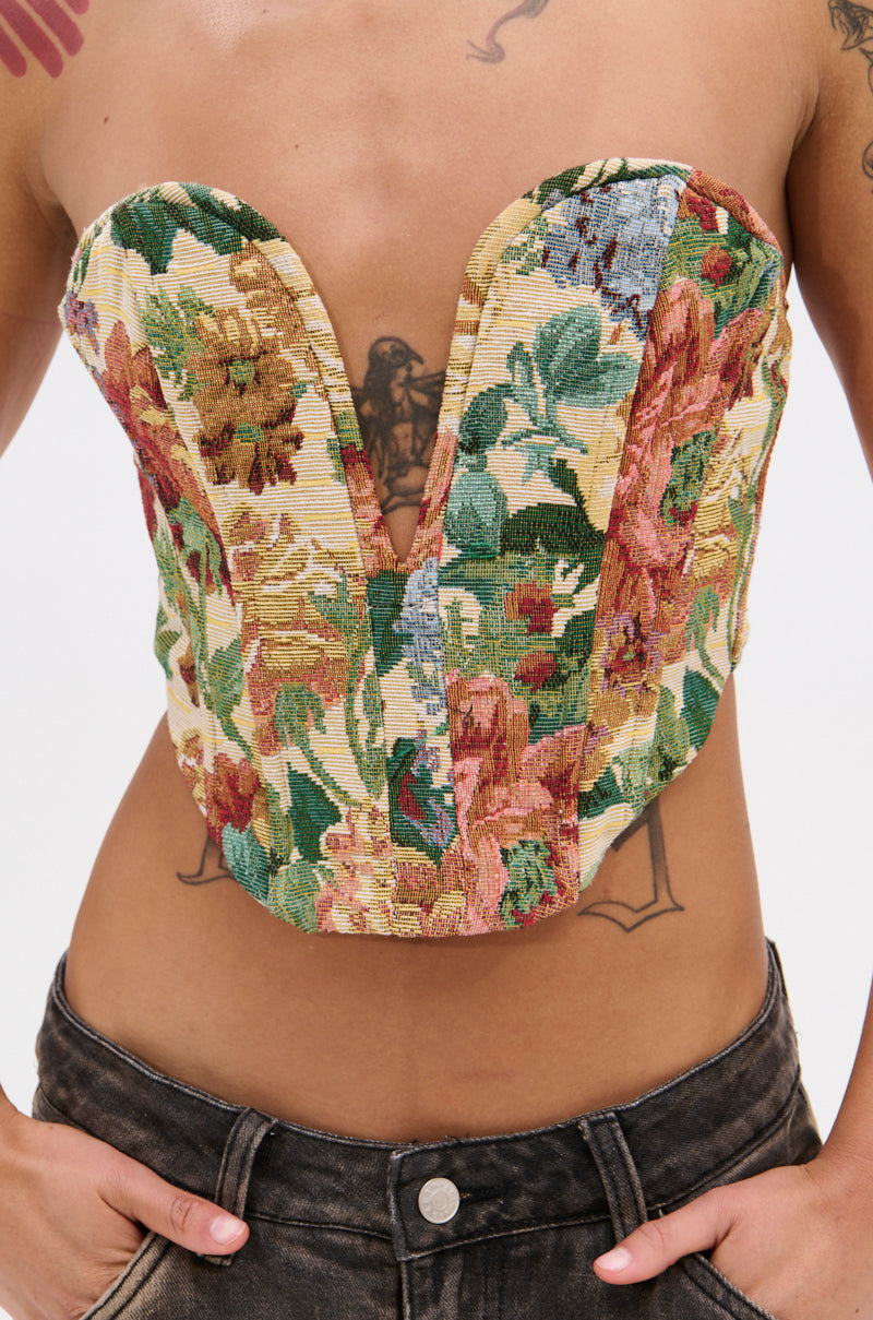 A person wears the BRIGETTE DISTRESSED JACQUARD CORSET featuring a strapless, deep V-cut neckline, paired with dark high-waisted jeans. Tattoos are visible on their chest and arms.