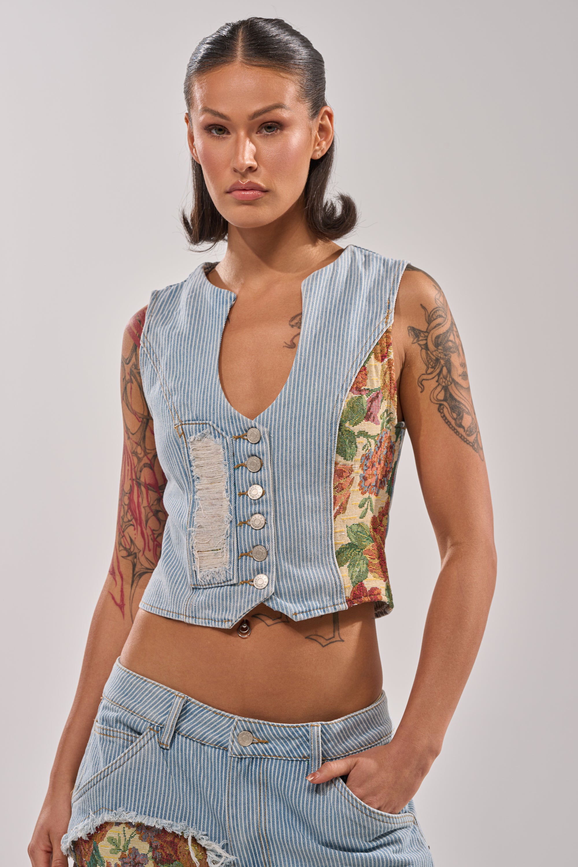 A woman with long dark hair and visible arm tattoos stands confidently, wearing the GUESS WHO STRIPE DENIM BUTTON DOWN with pinstripes, vintage floral panels, and matching distressed denim shorts. 0