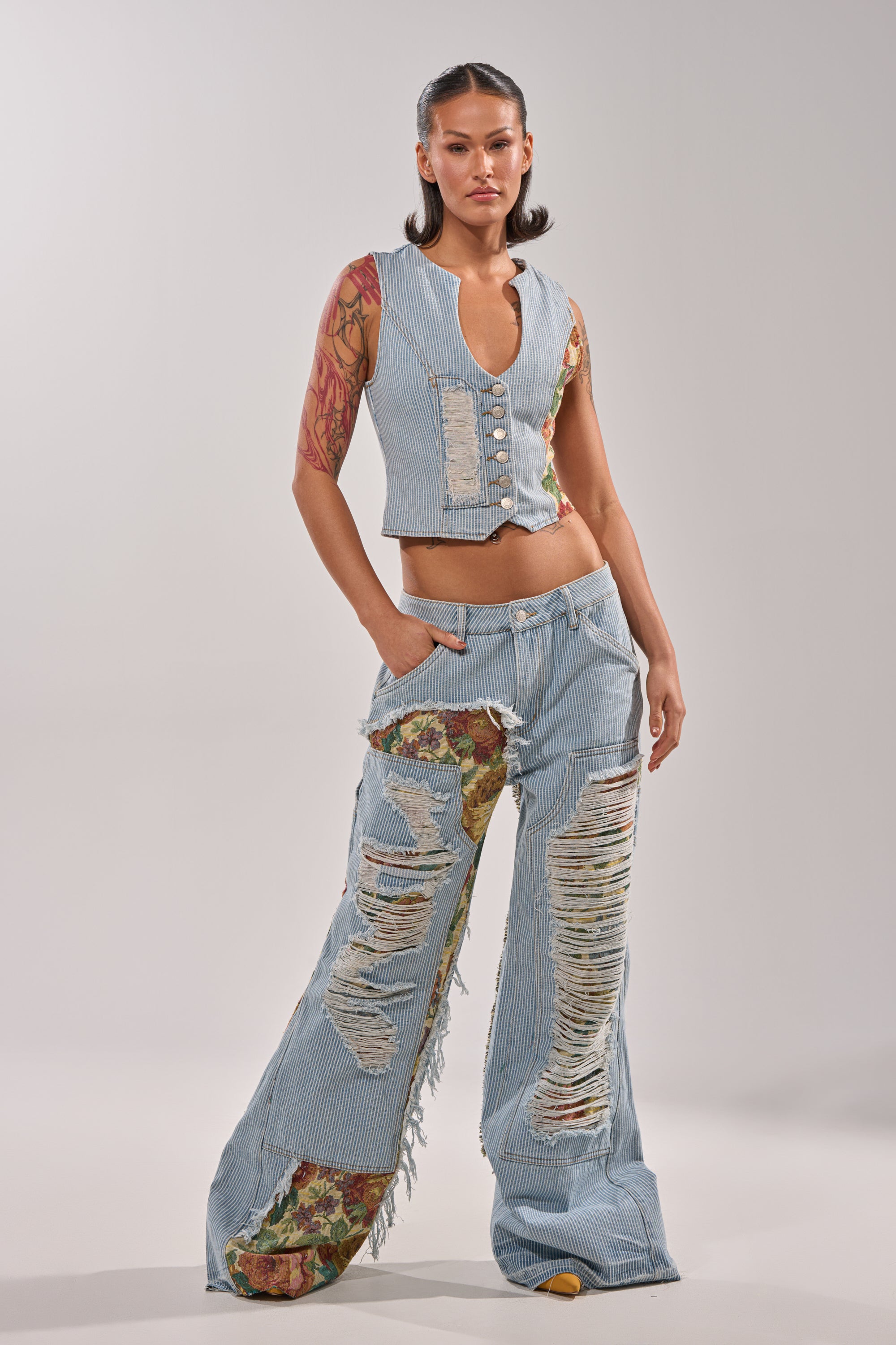 A woman with tattoos and slicked-back hair stands confidently in the GUESS WHO STRIPE DENIM BUTTON DOWN vest, styled with wide-leg jeans that feature distressed rips and vintage floral brocade patches. 5