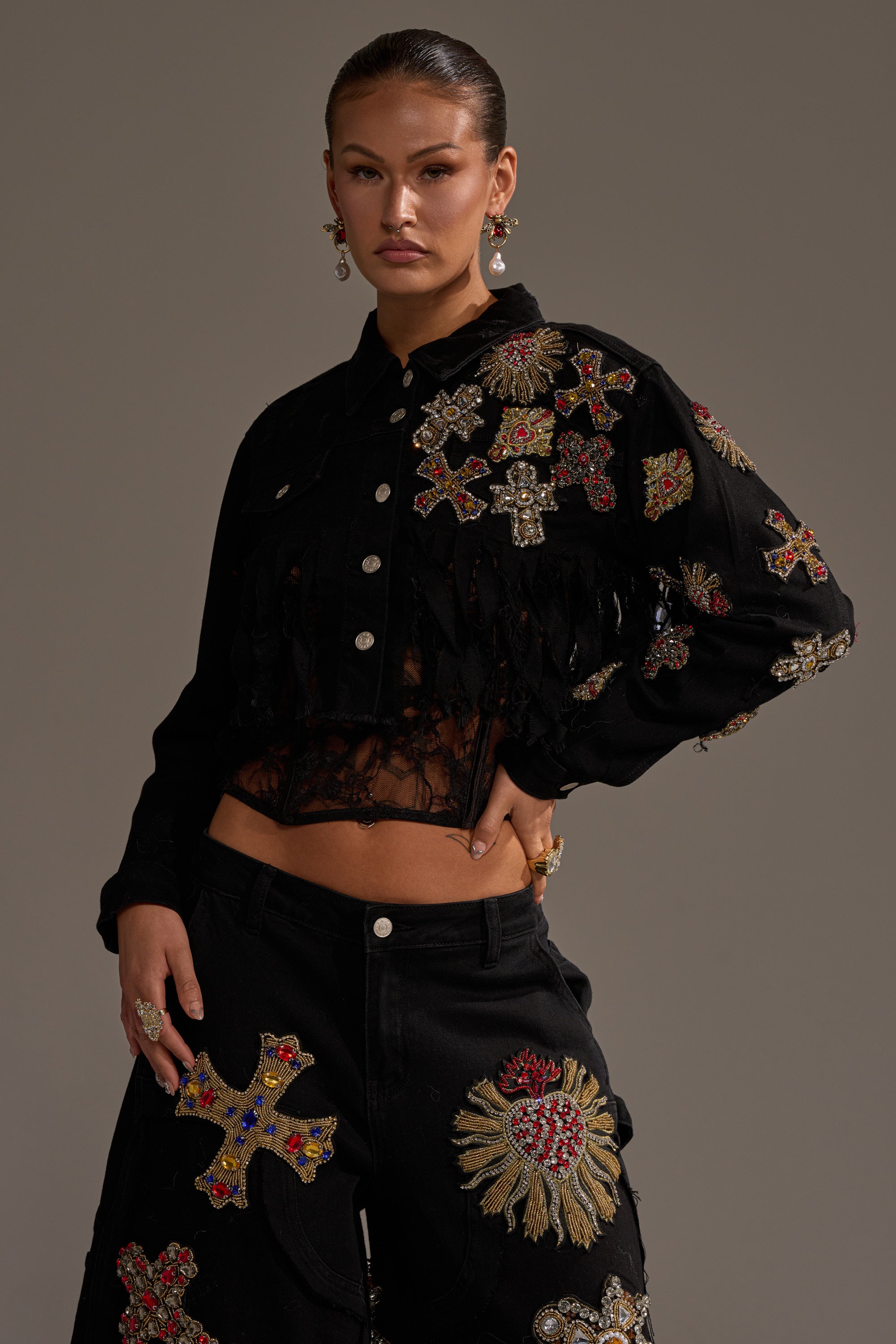 A woman poses in the MAIN SQUEEZE CROPPED DENIM JACKET, detailed with colorful cross and sunburst embellishments. She stands with one hand on her hip, wearing a neutral expression against a plain gray backdrop. 0