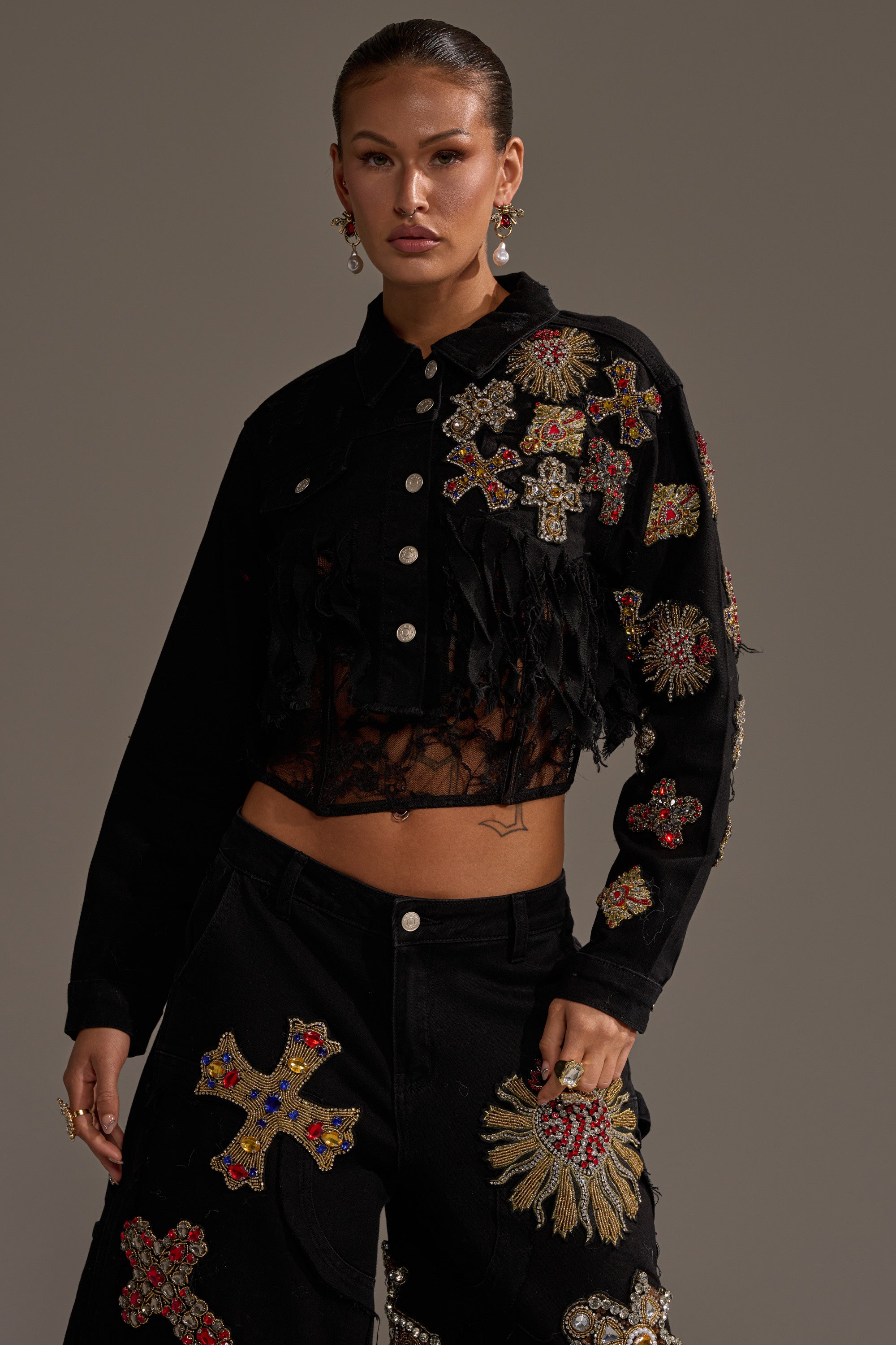 A woman stands confidently in the MAIN SQUEEZE CROPPED DENIM JACKET, featuring intricate, colorful beaded cross and sunburst designs. Her hair is pulled back, she wears dangling earrings, and the background is neutral. 2