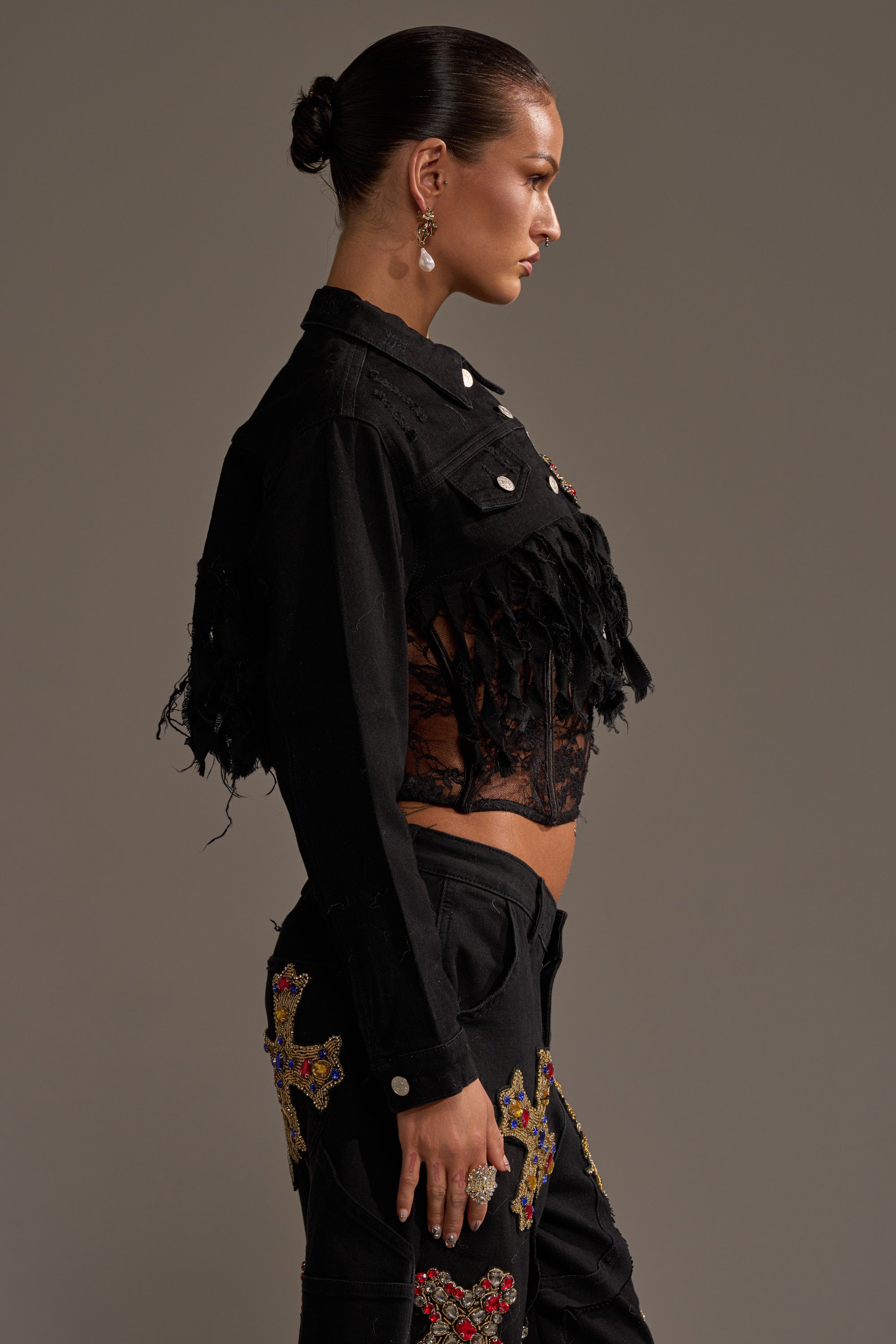 A woman stands in profile against a plain background, wearing the MAIN SQUEEZE CROPPED DENIM JACKET featuring distressed fringe and lace details, styled with black pants decorated with colorful embroidered crosses. Her hair is slicked back in a bun. 3