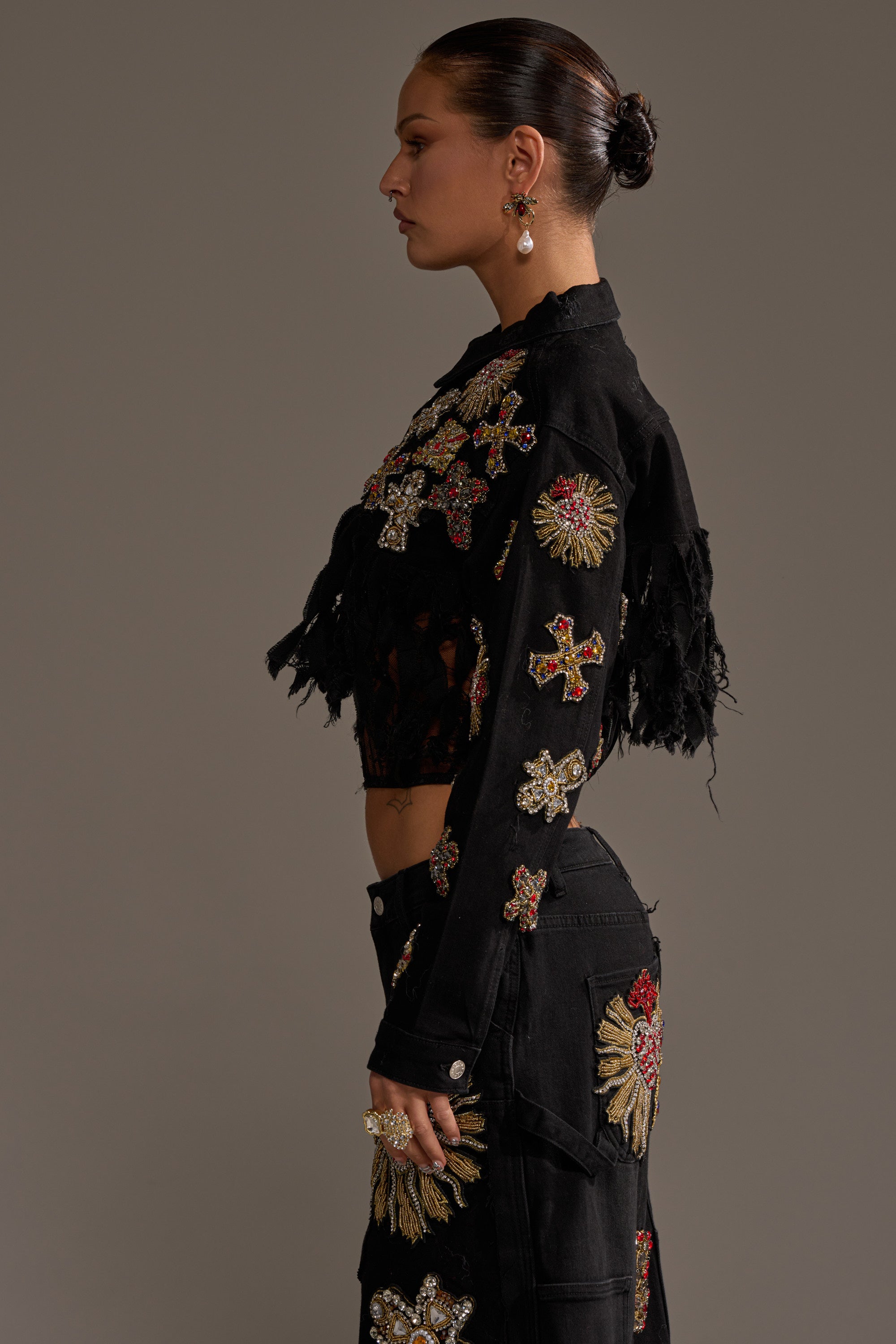 A woman with slicked-back hair models the MAIN SQUEEZE CROPPED DENIM JACKET and matching pants, both black and adorned with colorful embroidered patches and beads, posing sideways against a plain background. 4