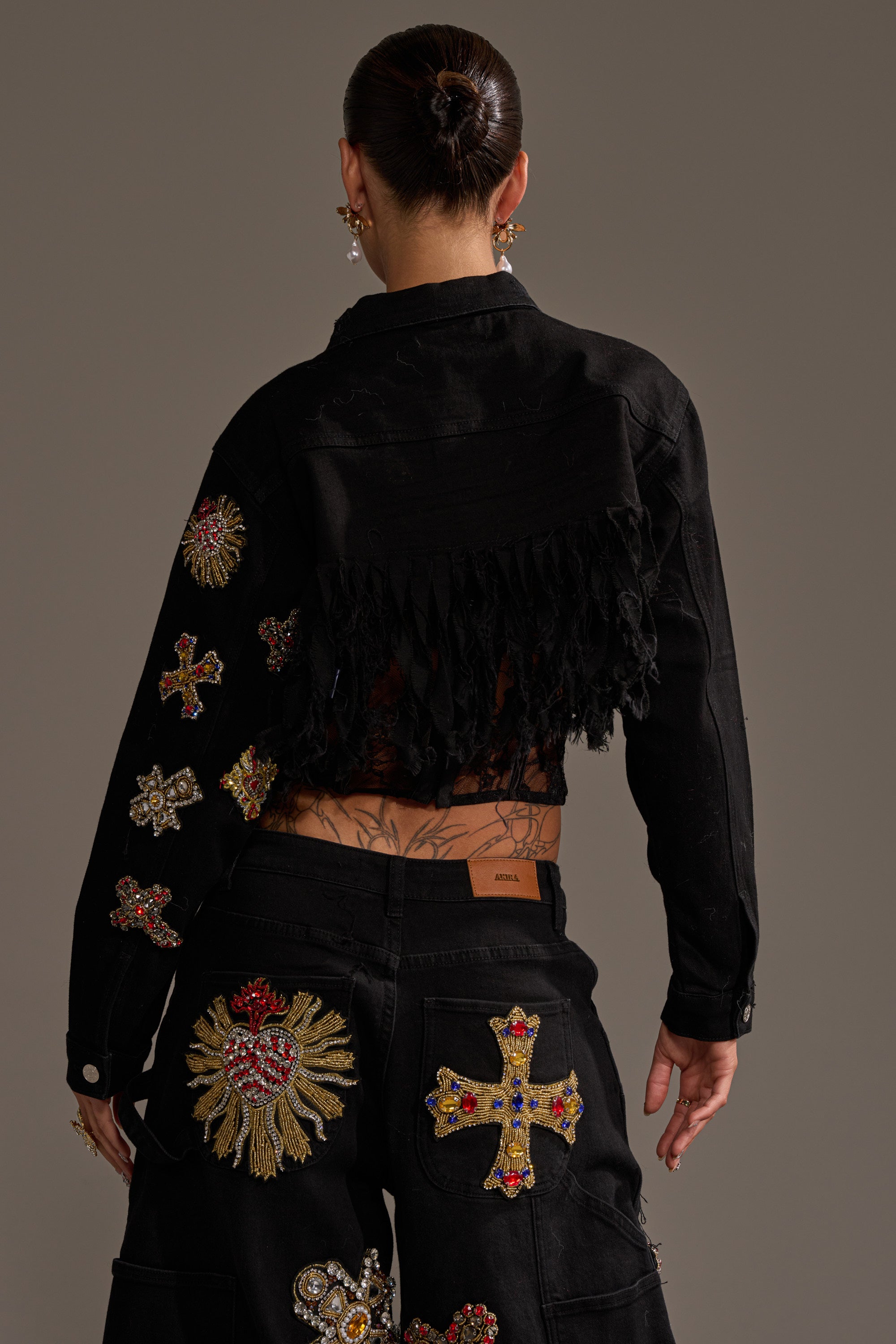 A person with dark hair in a bun, seen from behind, wears the MAIN SQUEEZE CROPPED DENIM JACKET—black with distressed fringe—paired with pants featuring colorful embroidered patches and beaded crosses, against a plain background. 5