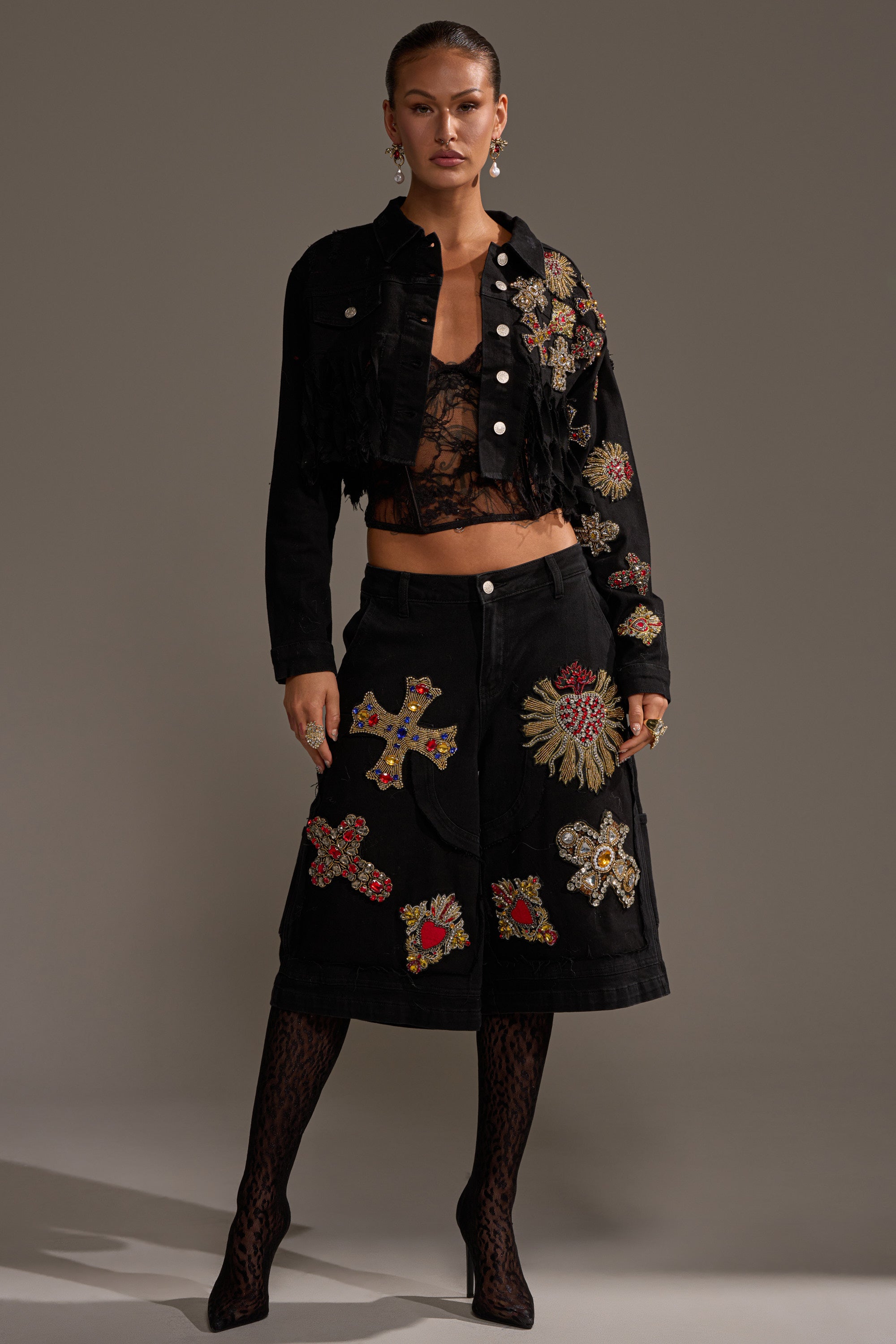 A woman confidently wears the MAIN SQUEEZE CROPPED DENIM JACKET, embellished with colorful embroidered patches like crosses and sunbursts, paired with a lace top, patterned tights, a matching skirt, and hoop earrings. 6
