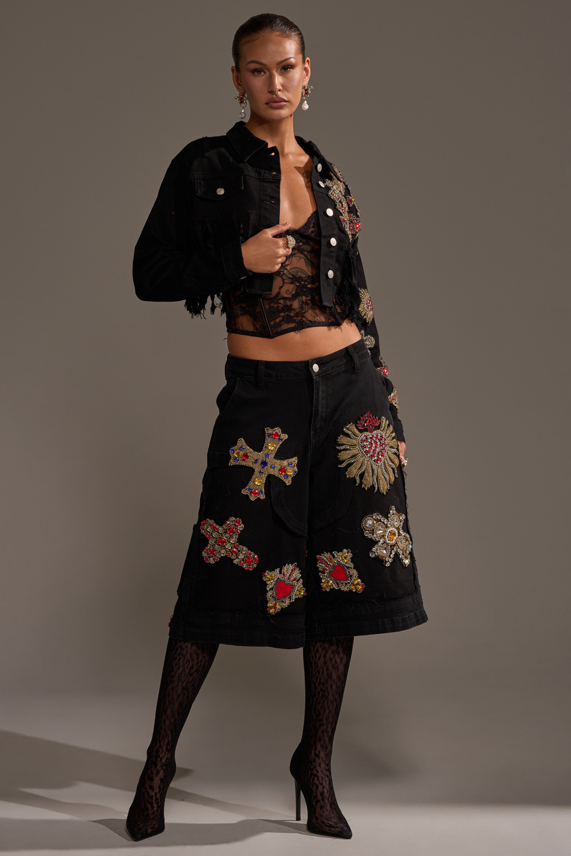 A woman poses confidently in the MAIN SQUEEZE CROPPED DENIM JACKET adorned with ornate cross-shaped patches, paired with a black lace top, distressed fringe denim skirt, patterned tights, and black heels against a neutral background. 7
