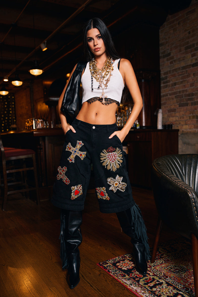 A woman with long dark hair stands confidently in a dimly lit bar, wearing a white crop top, layered necklaces, a black jacket over her shoulder, and the WILD AND FREE DENIM SHORTS.