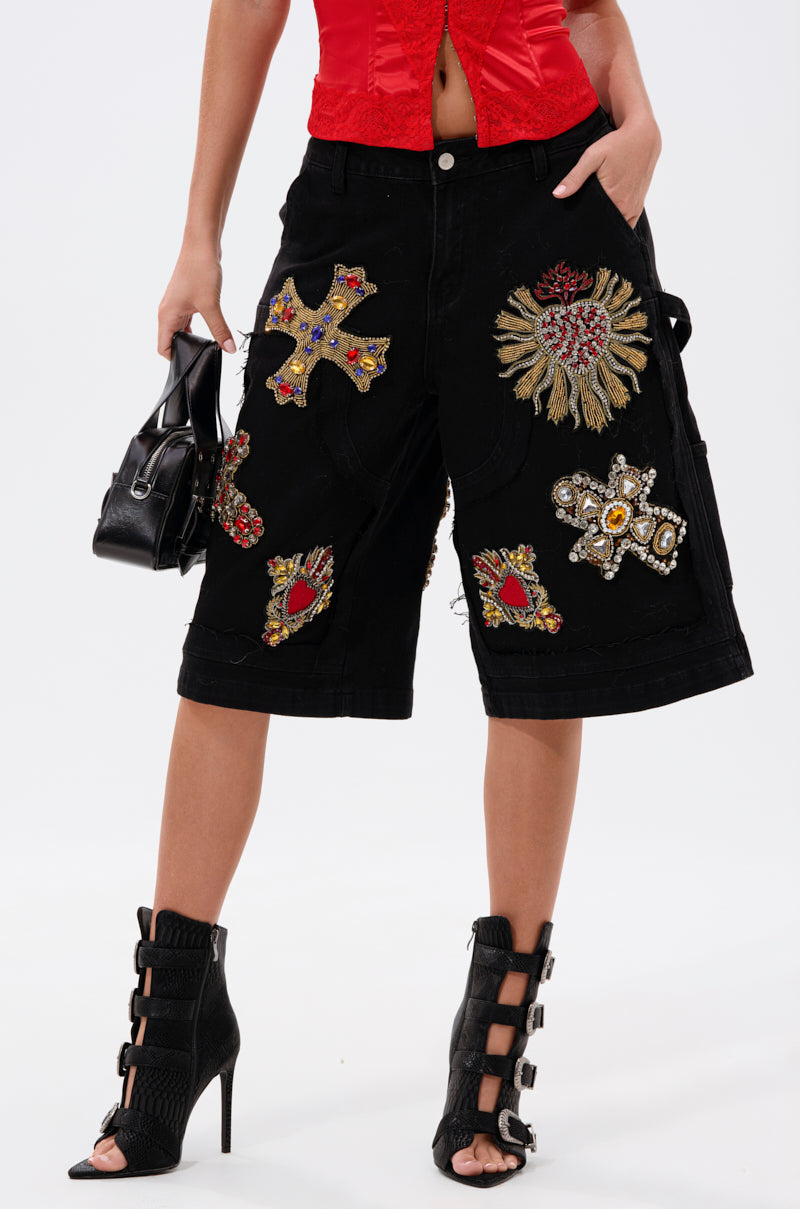 A person wears WILD AND FREE DENIM SHORTS—black, baggy bermudas with colorful embroidered patches—paired with a red top, black high-heeled buckled sandals, and a small black handbag.