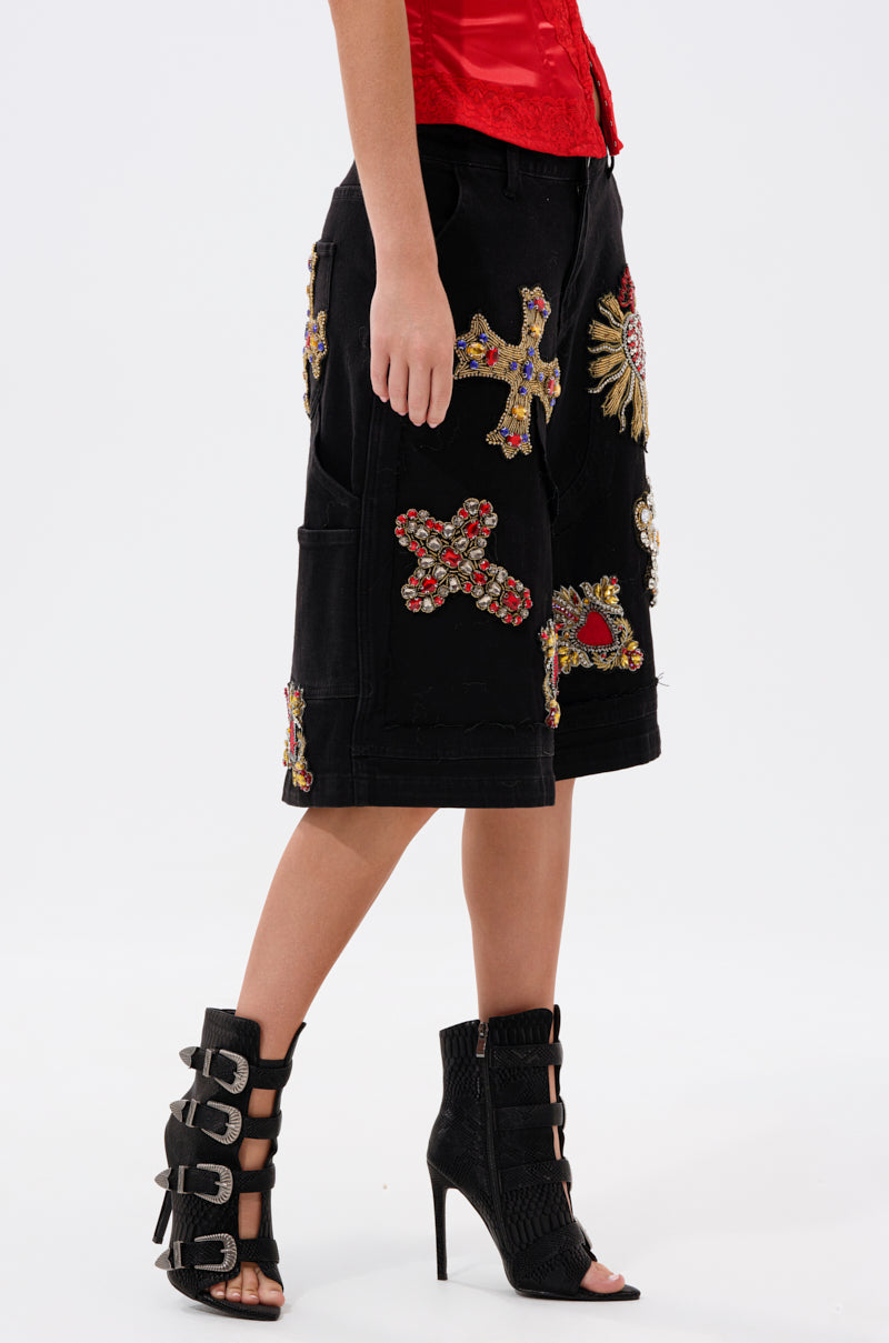 A person models the WILD AND FREE DENIM SHORTS—black, baggy bermuda shorts with ornate embroidered patches and crosses—paired with a red top and black high-heeled buckle booties, standing against a plain white background.
