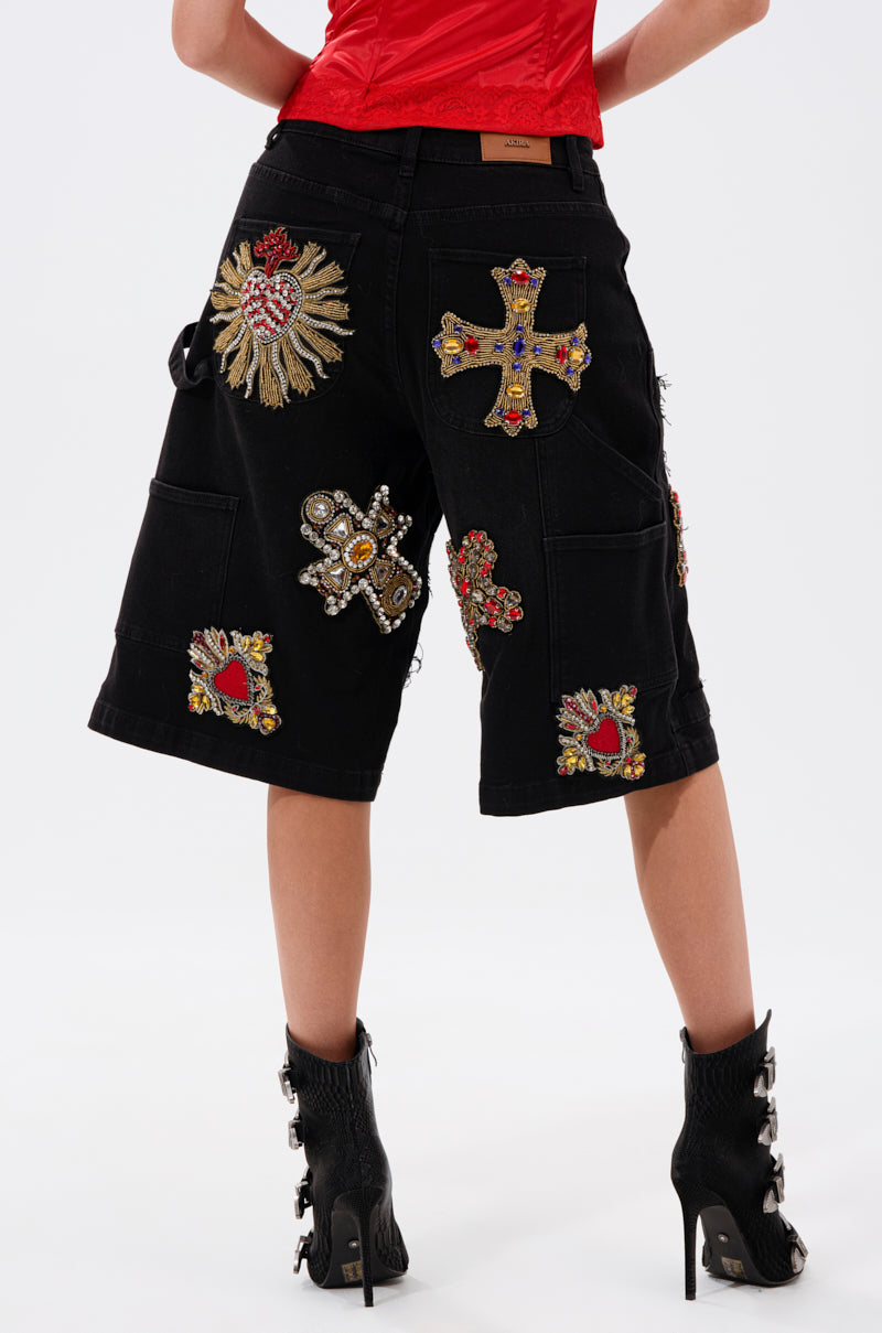 A person wearing WILD AND FREE DENIM SHORTS with cross and heart patches, a red top, and black heeled boots with buckles is shown from the back against a plain background.
