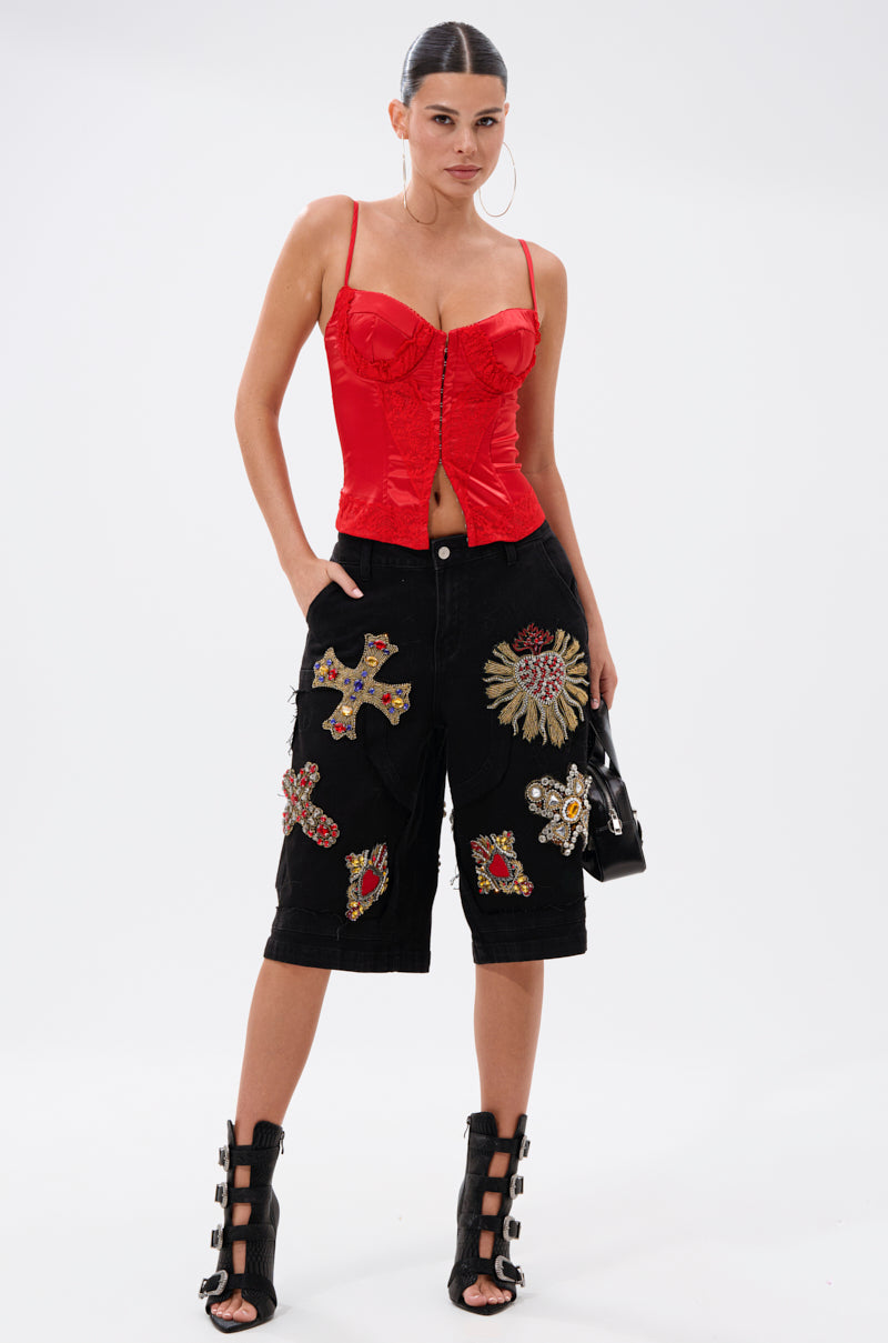 A woman stands confidently in a red corset top, WILD AND FREE DENIM SHORTS with colorful patches, black high-heeled sandals, large hoop earrings, and holds a small black bag.