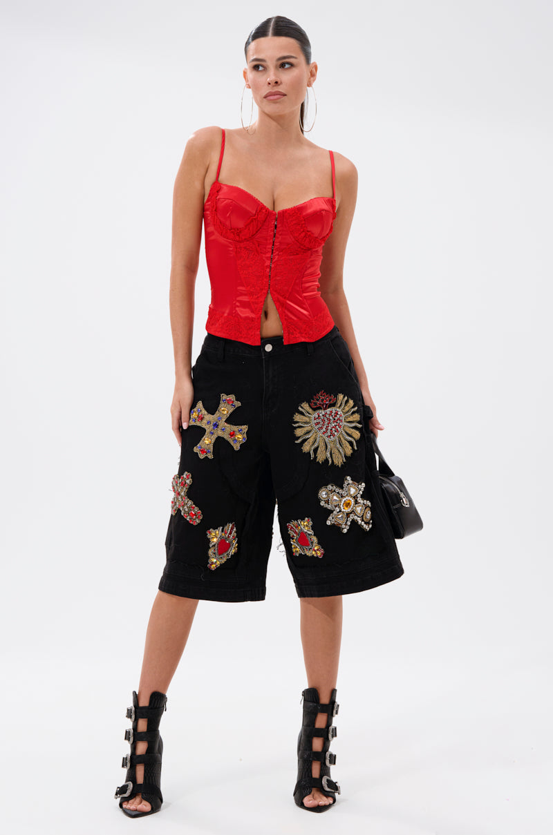 A woman poses against a white background, wearing a fitted red corset top, WILD AND FREE DENIM SHORTS with jeweled patches, black strappy heeled sandals, hoop earrings, and holding a black handbag.