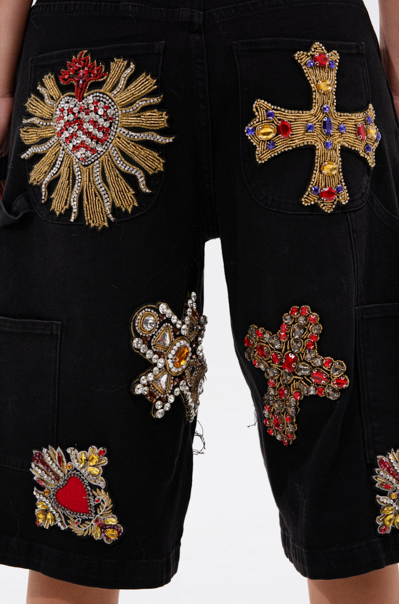 A close-up of the WILD AND FREE DENIM SHORTS shows black baggy bermuda shorts with ornate gold, red, and silver heart and cross embroidered and beaded patches on the back and sides for standout style.