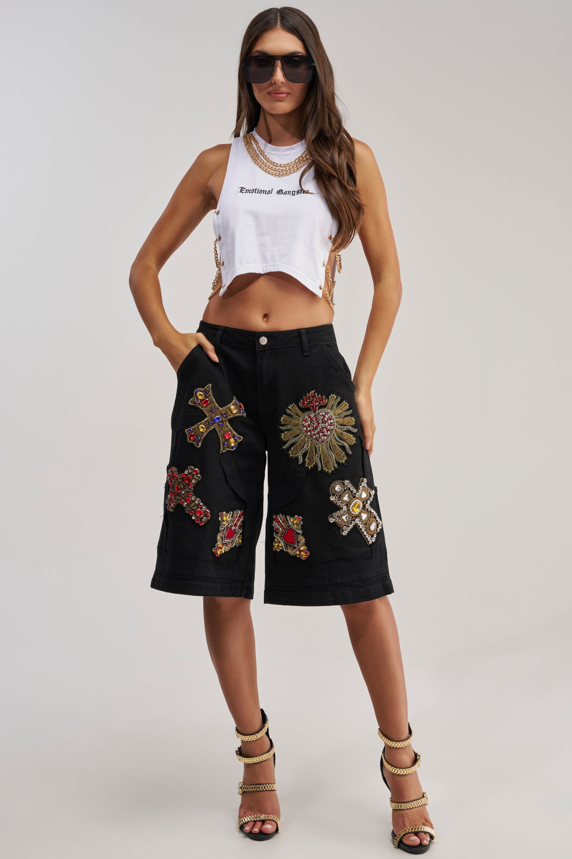A woman stands confidently in WILD AND FREE DENIM SHORTS—black bermuda shorts with large, colorful embroidered patches—paired with a white "Emotional Gangster" cropped tank top, sunglasses, and high heels, hands in her pockets.