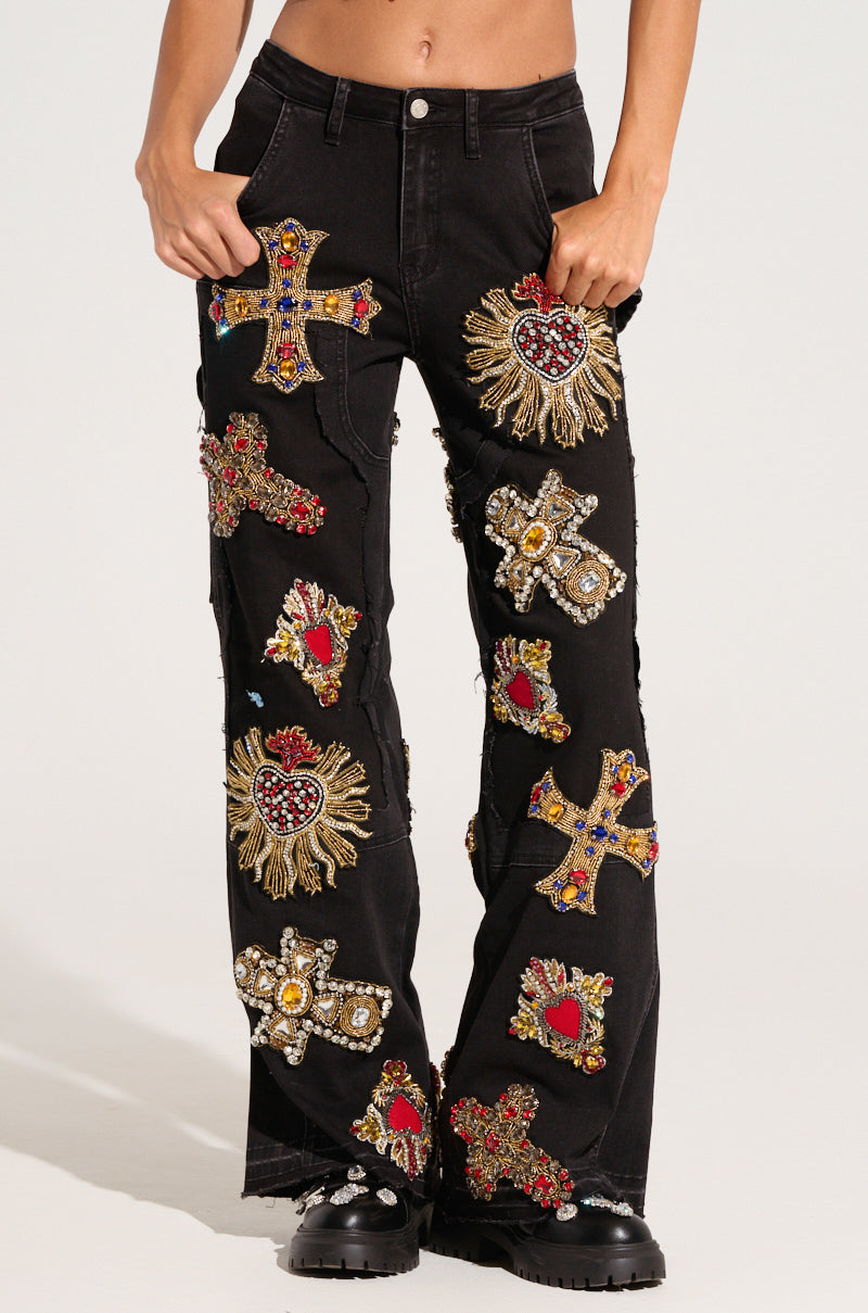 A person dressed in dark wash denim and black boots showcases the WILD AND FREE DENIM PANT, distinguished by numerous embroidered cross and heart patches embellished with beads and fringe details. From a waist-down perspective, their hands are casually placed in the pants' pockets.