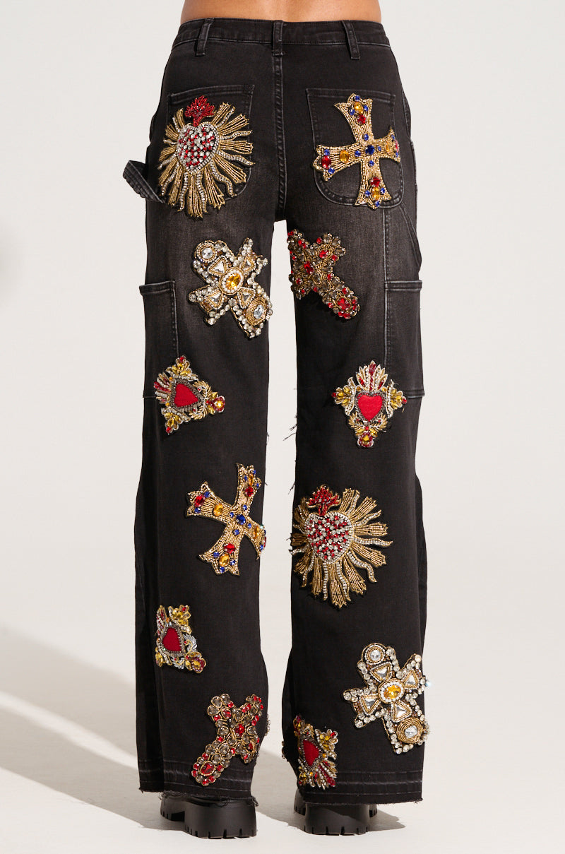 A rear view shows a person wearing the WILD AND FREE DENIM PANT, embellished with vibrant, ornate patches showcasing embroidered crosses and heart designs. The patches are meticulously adorned with beads and embroidery on dark wash denim. The individual stands on a light-colored floor, captured from the waist down.