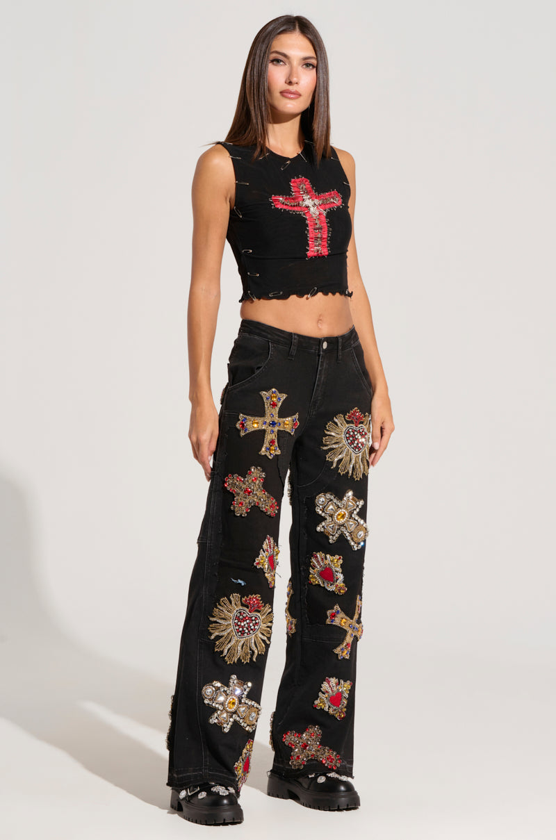 A woman stands in a studio wearing a black crop top with a large embellished red cross and WILD AND FREE DENIM PANT adorned with various colorful, embroidered cross and heart-shaped patches. She is also wearing black platform boots and has straight brown hair.