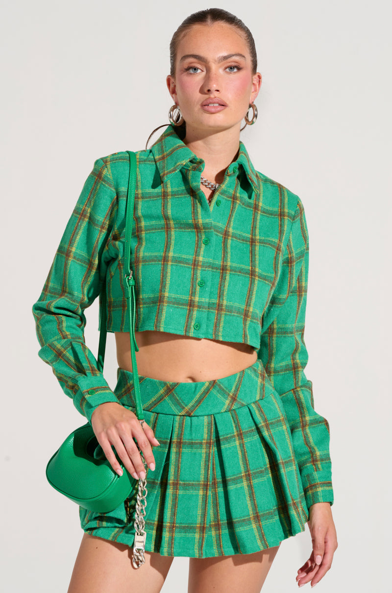 A person wearing a green plaid outfit is sporting the LUCKY GIRL BUTTON DOWN SHIRT, which has a cropped boxy silhouette, paired with a pleated mini skirt. They are holding a green handbag with a chain strap and accessorized with hoop earrings and a necklace while standing against a plain background.