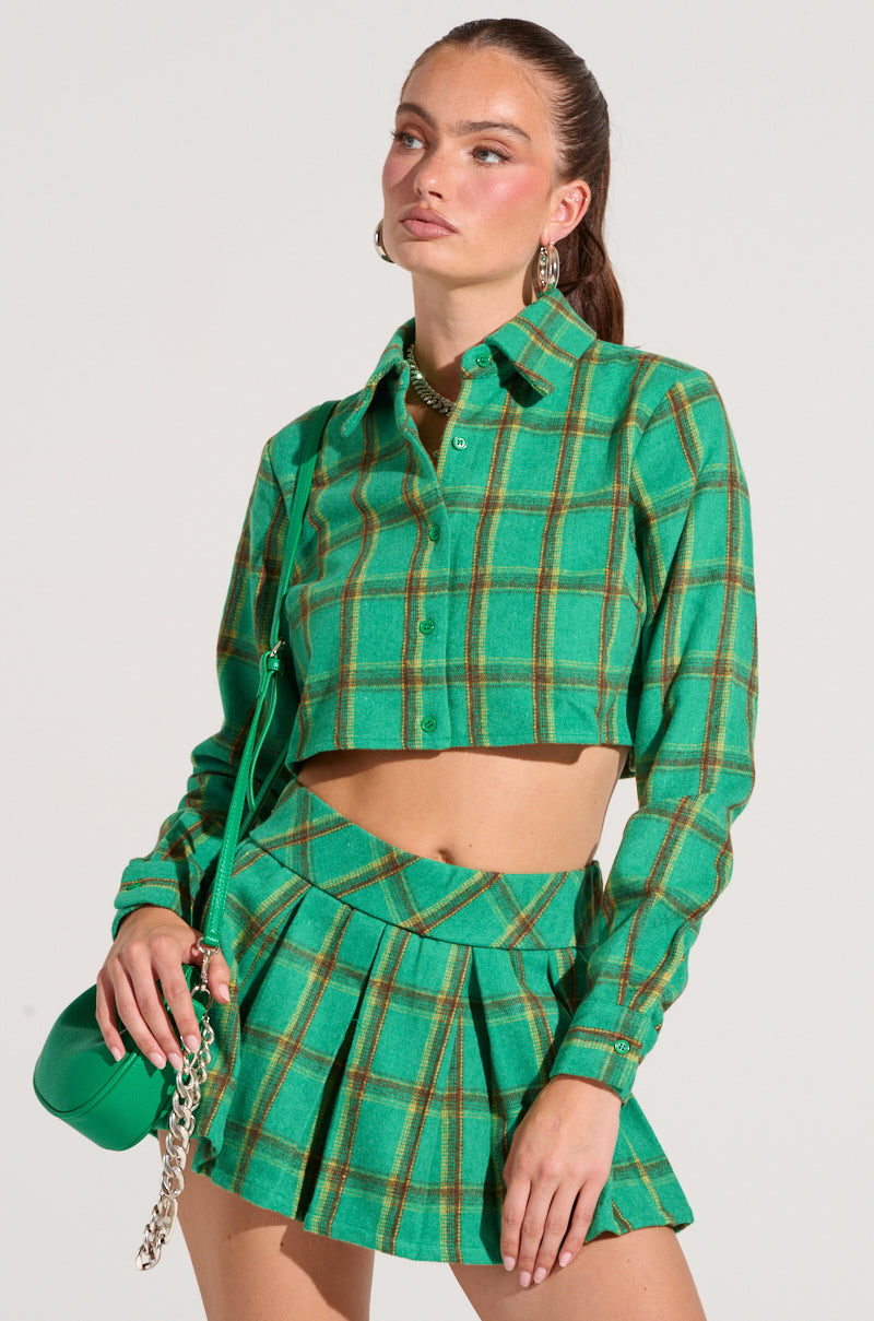 A person poses confidently wearing the Lucky Girl Button Down Shirt in green plaid and a matching pleated skirt, flaunting a cropped boxy silhouette. They hold a matching green bag with a chain strap, their hair pulled back and accessorized with earrings. The backdrop is simple and neutral, highlighting the playful flannel fabrication of this ensemble.