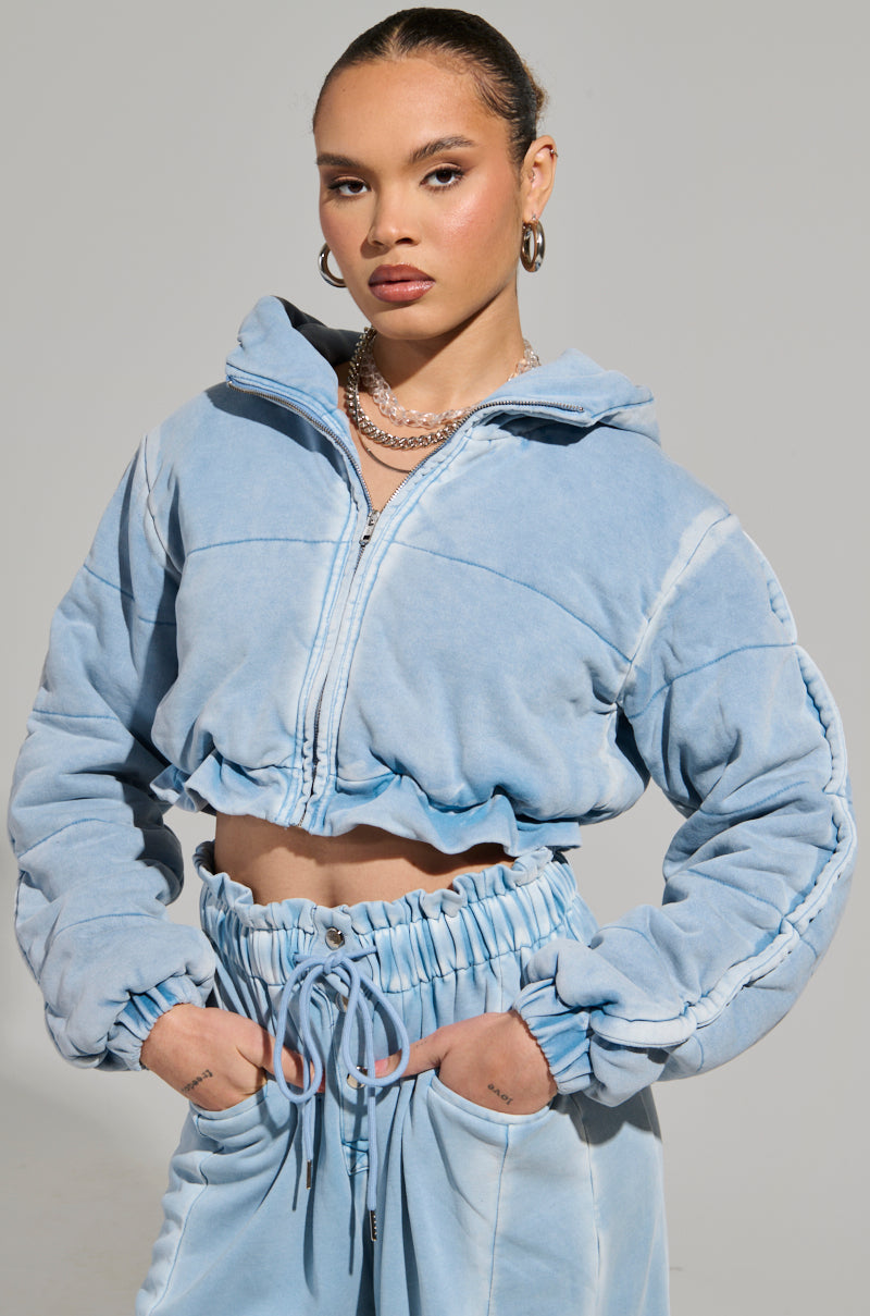A person with tied-back hair is sporting the HONEY HONEY FRENCH TERRY BOMBER in a light blue acid wash, paired with matching high-waisted sweatpants. They stand confidently with their hands in their pockets against a plain background, accessorized with hoop earrings and a chain necklace.