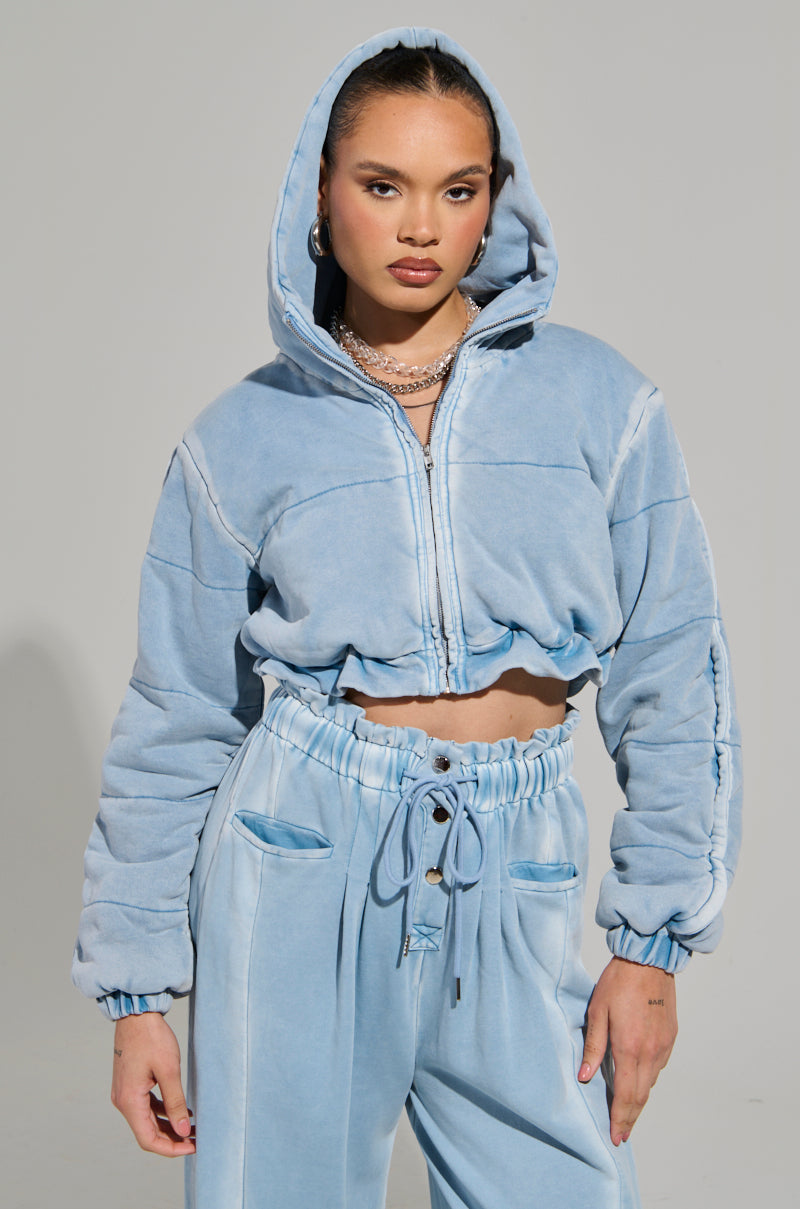 A person wearing the HONEY HONEY FRENCH TERRY BOMBER in light blue velvet and matching pants poses confidently. The cropped fit of the bomber, complete with a zipper and hood up, enhances their style, while the pants feature a drawstring waist. A plain, neutral background highlights their chic acid wash look.