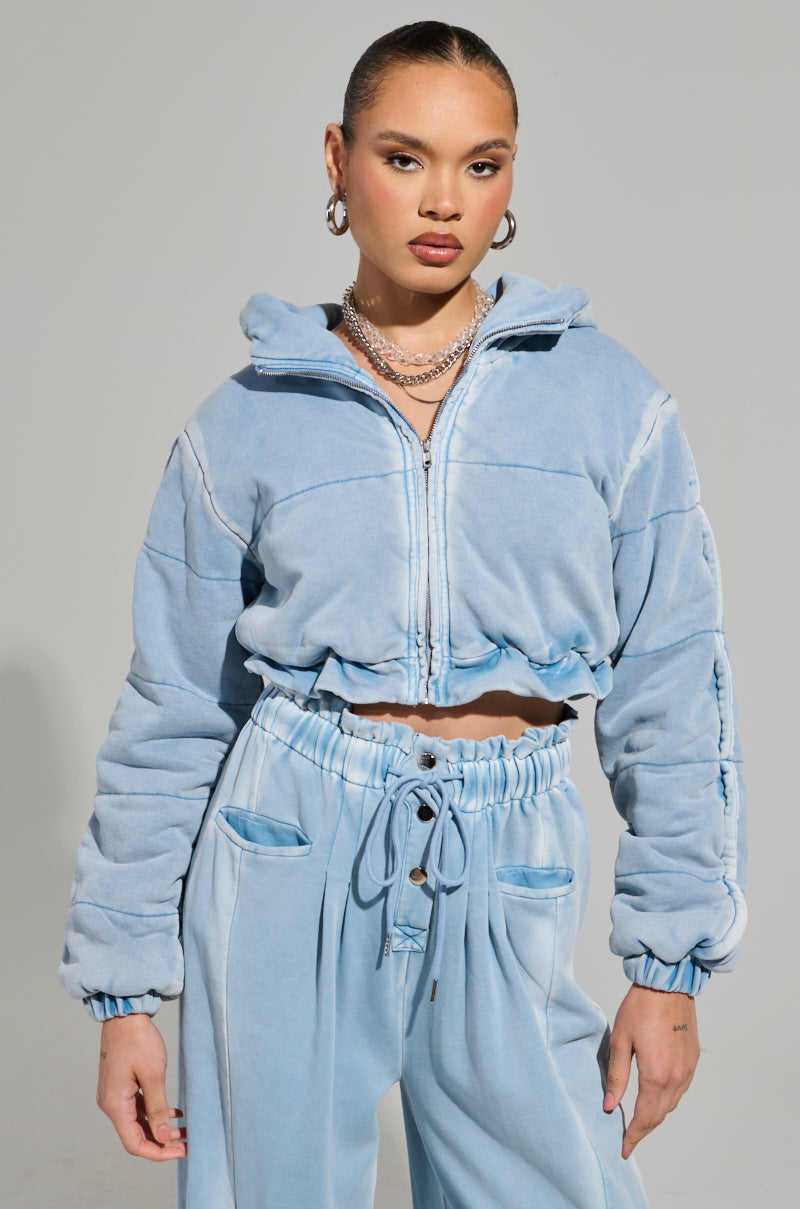 A person is dressed in the HONEY HONEY FRENCH TERRY BOMBER, a light blue, plush zip-up jacket paired with matching jogger pants that feature a cropped fit. Their hair is styled in a sleek bun, complemented by layered necklaces and hoop earrings. The setting is a plain, light gray background.