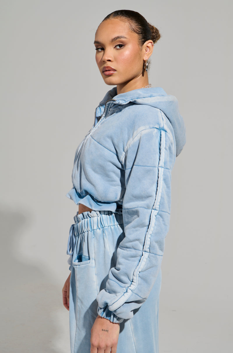 A person with tied-back hair poses confidently in the HONEY HONEY FRENCH TERRY BOMBER, showcasing its light blue, cropped fit. They are angled slightly to the side with a neutral expression against a plain gray backdrop, embodying the casual coolness of an acid wash finish.