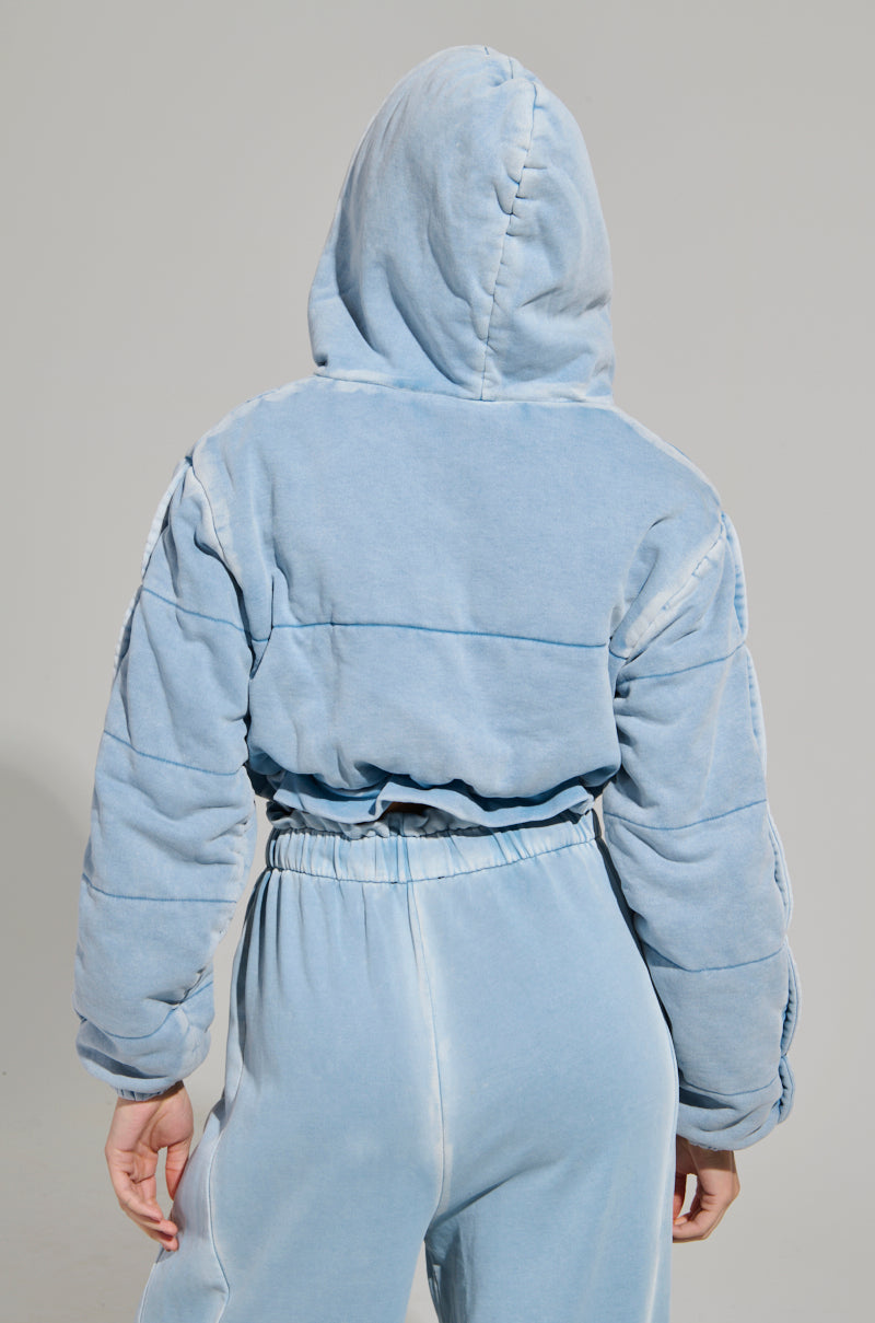 A person wearing the HONEY HONEY FRENCH TERRY BOMBER in light blue, styled with matching cropped-fit pants. The hood is up, covering their head as they stand against a plain background, facing away from the camera.