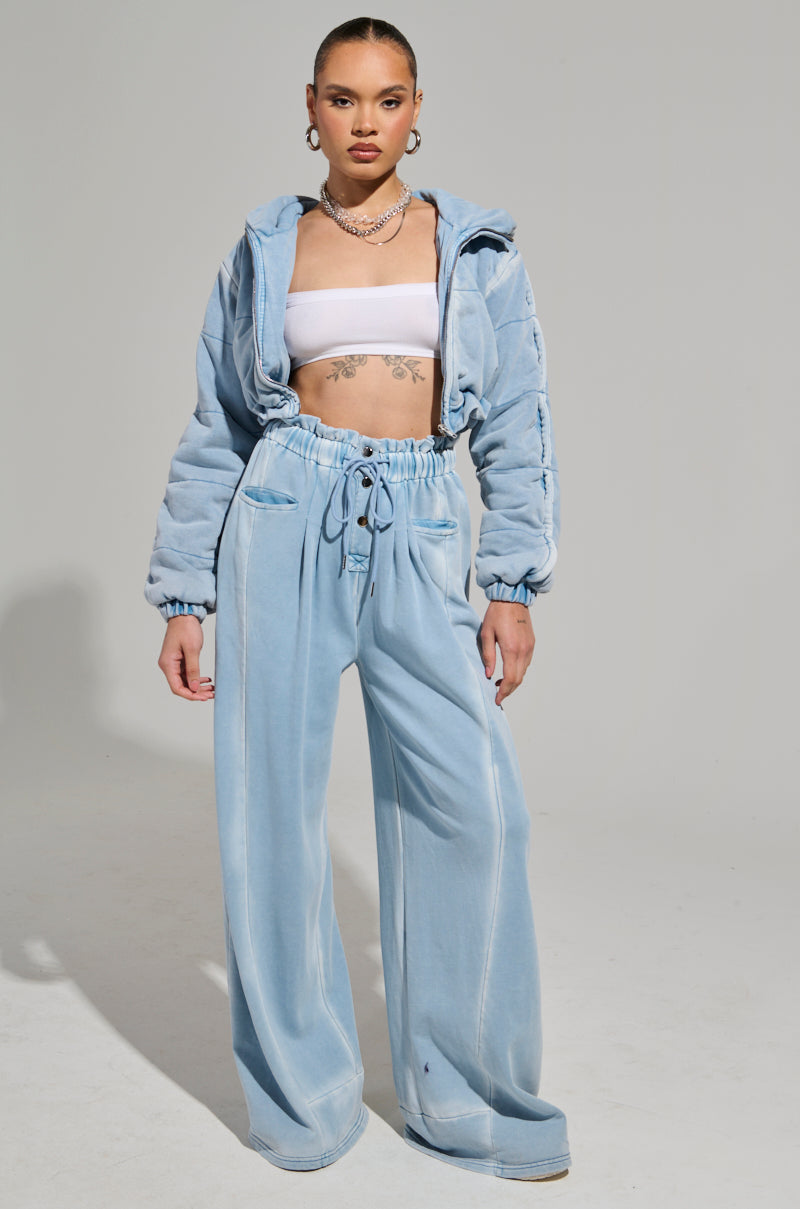 A person stands confidently dressed in the HONEY HONEY FRENCH TERRY BOMBER, a light blue velour tracksuit with wide-legged pants and a zip-up hoodie. Underneath, they sport a cropped fit white tube top. Accessories include large hoop earrings and layered necklaces against a plain gray background.