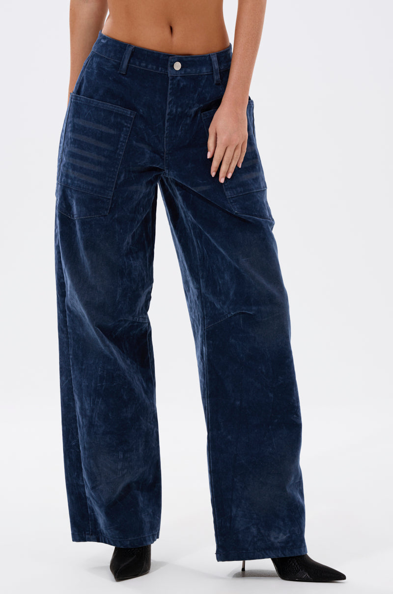 Someone wears the VALERIE VELVET EFFECT WIDE LEG PANT IN BLUE, featuring a high-rise waist and utility-style pockets, paired with black pointed-toe boots, standing against a plain white background.