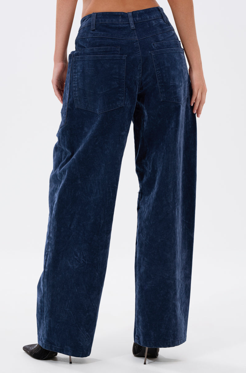 A person stands against a white background, wearing the VALERIE VELVET EFFECT WIDE LEG PANT IN BLUE with a high-rise waist and black heels. The photo is taken from behind, showcasing the pants’ utility-style pockets.