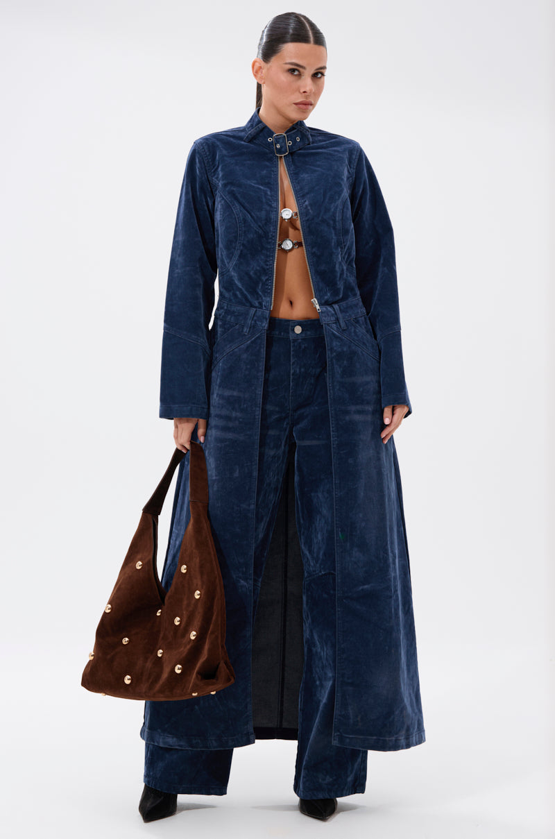 A woman models the VALERIE VELVET EFFECT WIDE LEG PANT IN BLUE with utility-style pockets, paired with a matching blue velvet coat and a brown bag with gold accents. Her hair is tied back as she poses against a white background.