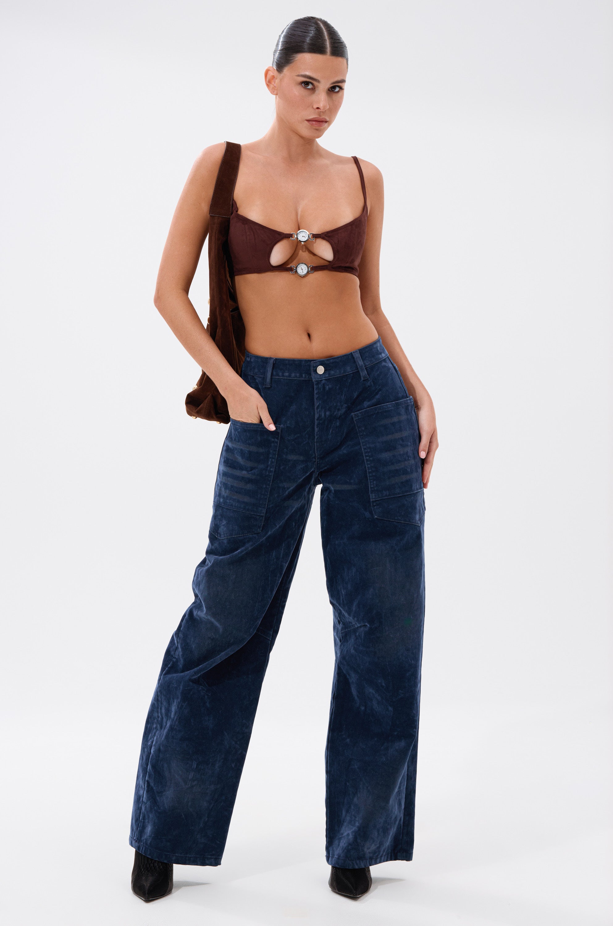 A woman poses confidently in the READ BETWEEN THE LINES CLOCK BRALETTE with cutouts and dark blue wide-leg pants, carrying a brown bag over her shoulder against a white background. 5