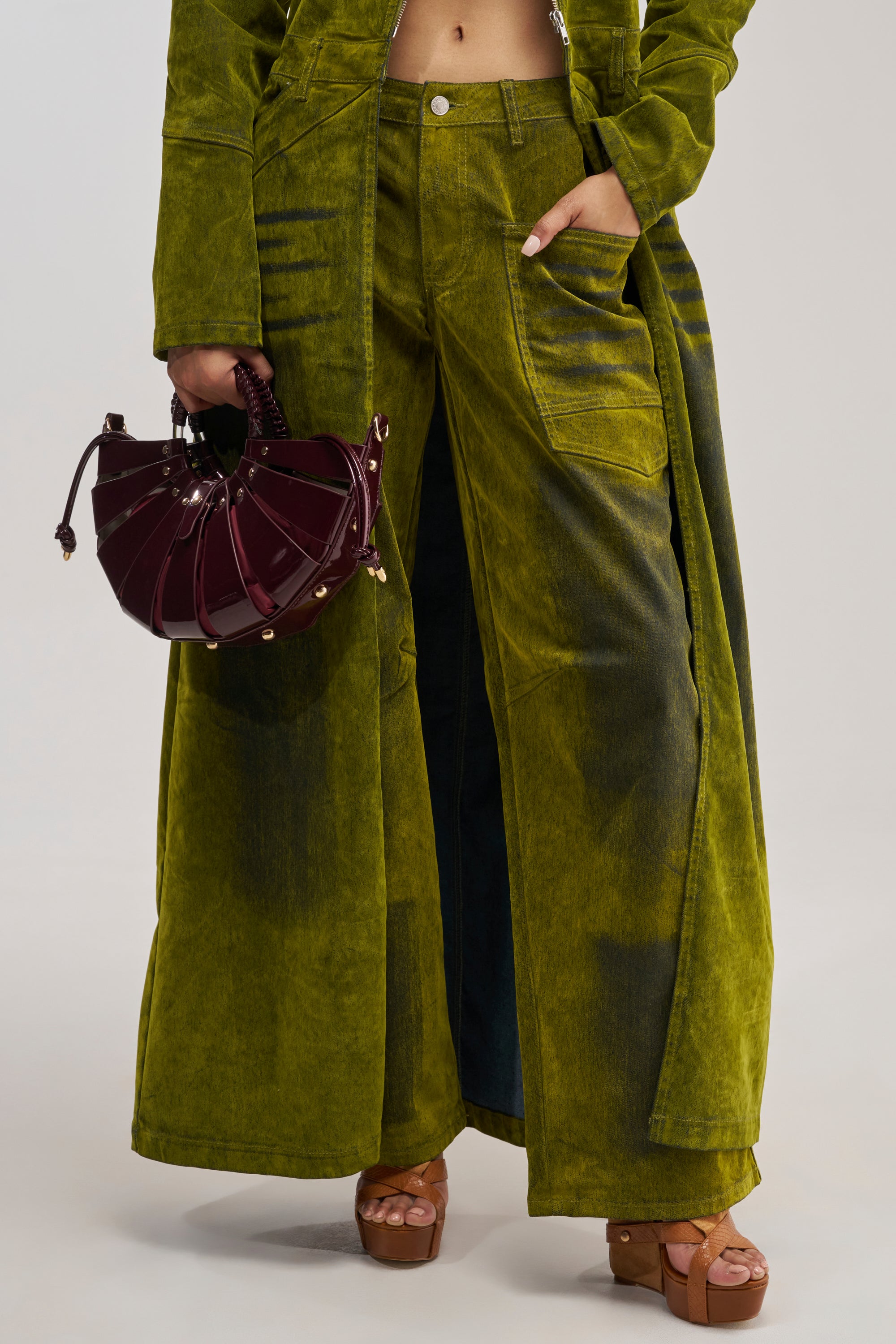 A person wearing the VALERIE VELVET EFFECT PANT IN GREEN with a matching coat holds a dark purple circular handbag. They are also wearing brown platform sandals. The background is plain and neutral.