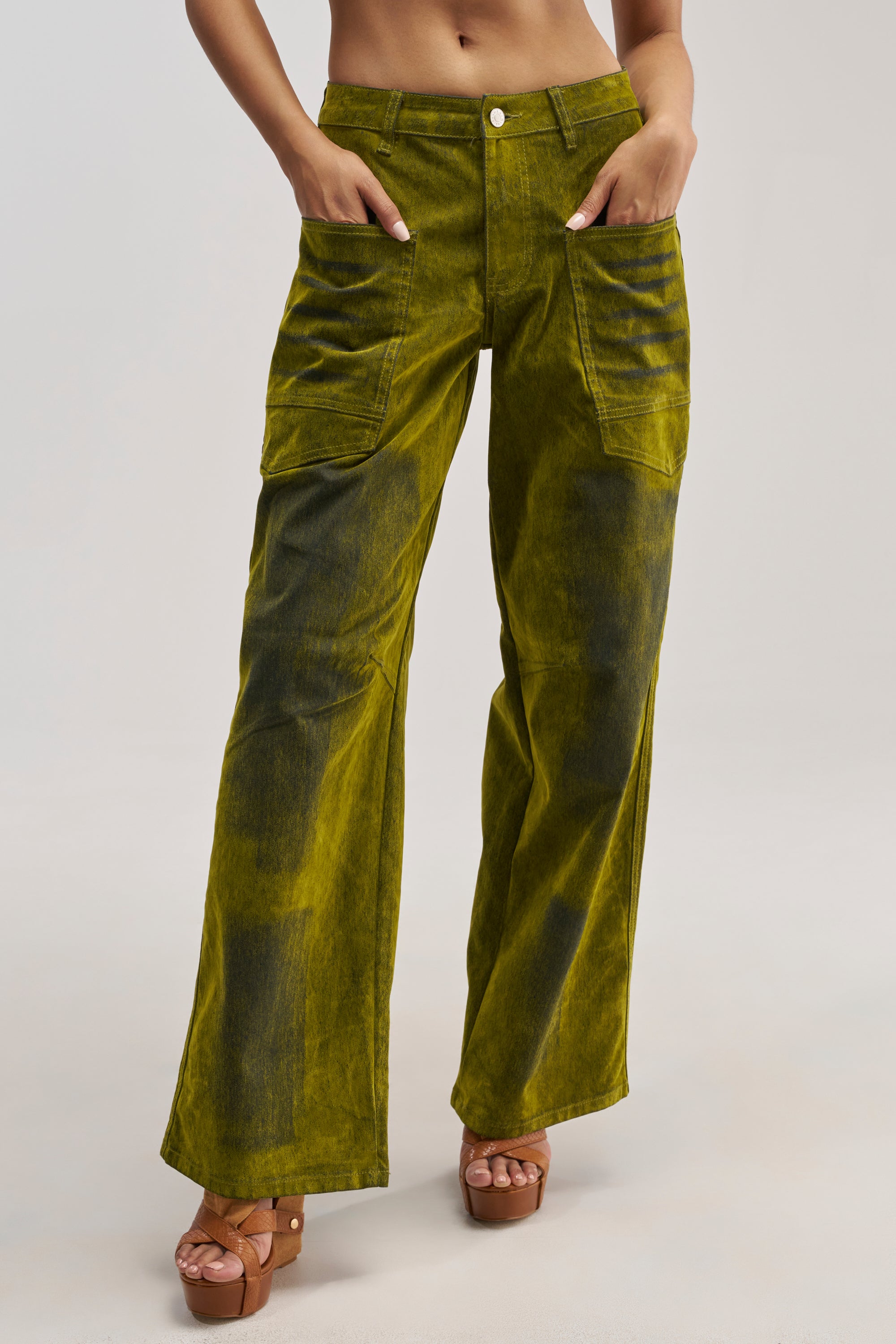 A person wearing the VALERIE VELVET EFFECT PANT IN GREEN—high-rise, wide-leg denim pants with a yellow-green velvet finish and large front pockets—paired with brown platform sandals, standing against a plain background.