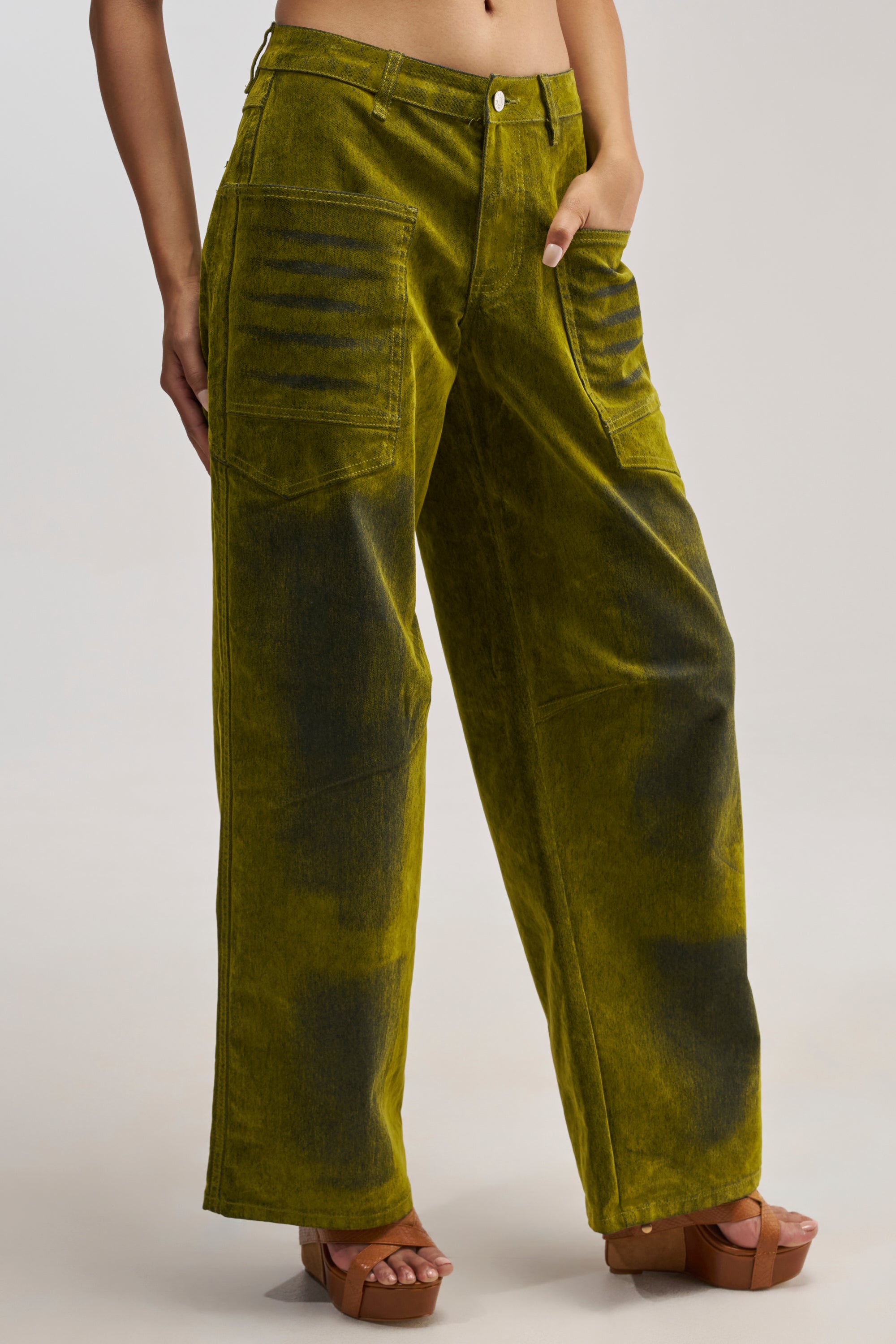 Wearing the VALERIE VELVET EFFECT PANT IN GREEN—high-rise, wide-leg olive green jeans with faded black patches and front pockets—paired with brown platform sandals; torso is out of frame.
