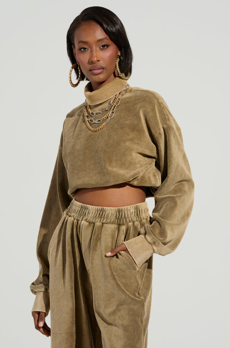 A woman wears a chic tan corduroy outfit, including the DRIFTING AWAY TOP by AKIRA Label paired with matching high-waisted pants. Adding cozy style to her look, she accessorizes with large hoop earrings and layered gold chains, posed against a neutral background.