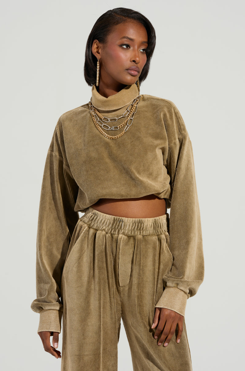 A person dressed in a cozy, beige corduroy outfit from the DRIFTING AWAY TOP collection by AKIRA Label poses against a neutral background. They are adorned with layered necklaces and hoop earrings, with their hair styled to the side, exuding an effortlessly chic vibe.