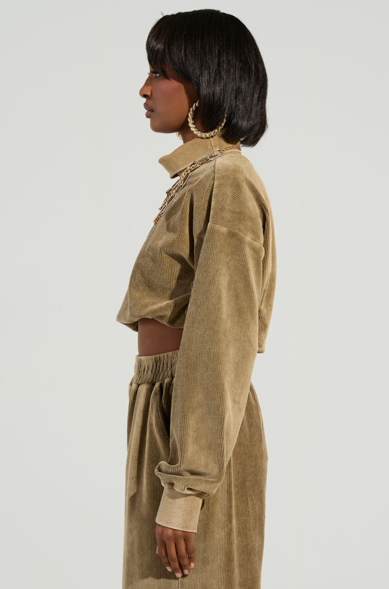 A person with shoulder-length dark hair stands in a cozy style, wearing a beige corduroy set that includes the DRIFTING AWAY TOP and high-waisted pants, accessorized with large hoop earrings against a neutral background.