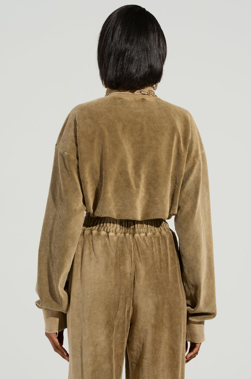 A person with shoulder-length dark hair is seen from the back, dressed in a brown corduroy tracksuit that radiates cozy style. The tracksuit, similar to the DRIFTING AWAY TOP by AKIRA Label, includes a long-sleeve top paired with matching pants. The neutral backdrop highlights the outfit's relaxed appeal.