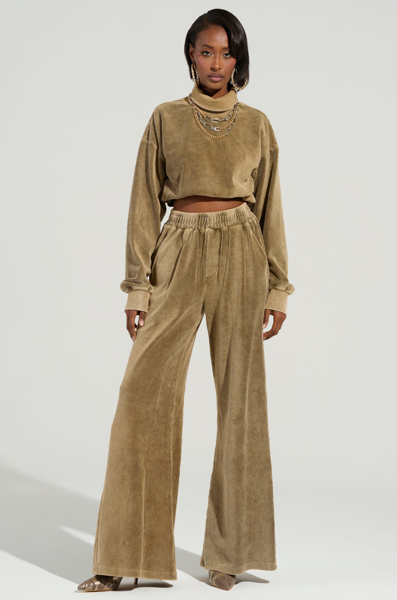 Dressed confidently in a cozy style, the person sports the ribbed knit beige velour DRIFTING AWAY TOP from the AKIRA Label collection. The ensemble includes a cropped, long-sleeve top paired with wide-leg pants, complemented by layered necklaces and peep-toe shoes against a plain background.