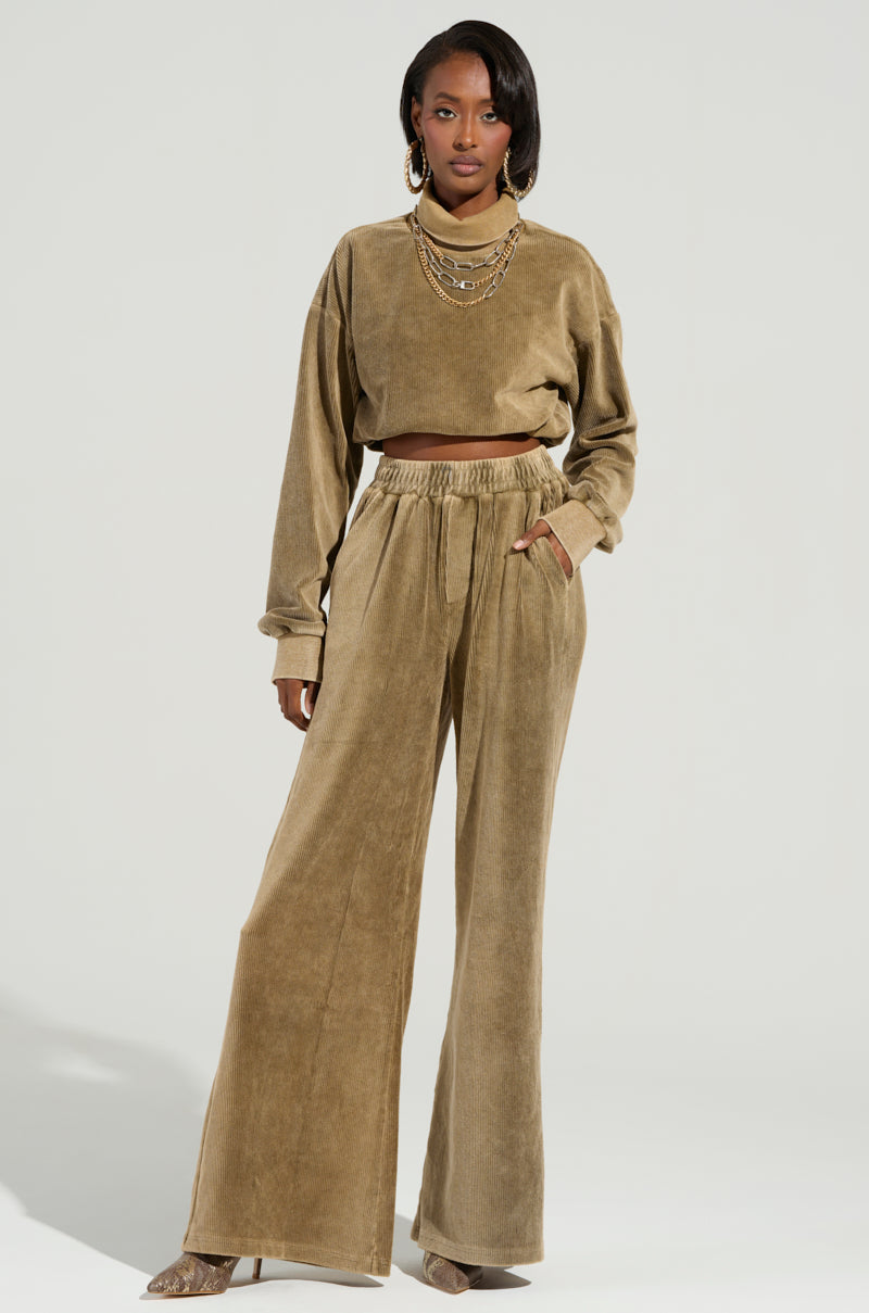 A person stands confidently in a stylish tan velvet two-piece, featuring the DRIFTING AWAY TOP by AKIRA Label and wide-leg pants. Accessorized with layered necklaces and earrings, they exude cozy style as they pose against a light gray background.