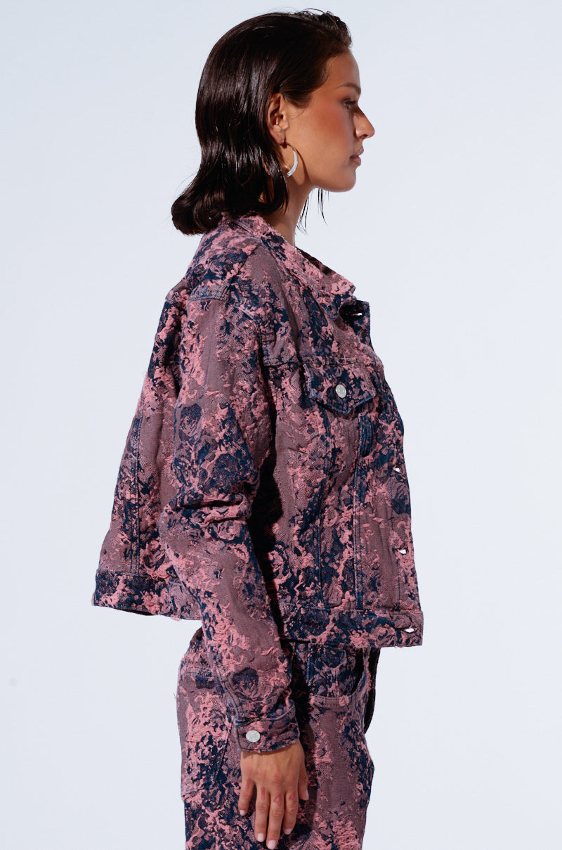 A person with short dark hair stands in profile against a plain background. They are wearing the JUST LIKE THAT DENIM JACKET and matching pants, both featuring an oversized fit with a pink and black marbled design. The jacket has buttons and front pockets, showcasing a distressed pink pattern. They are also wearing large hoop earrings.