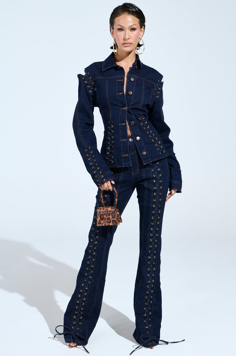 A person poses in a dark denim outfit featuring lace-up details on the jacket and high-rise, THATS ON ME FLARE DENIM PANT. They're holding a small leopard print handbag against a plain, light background.