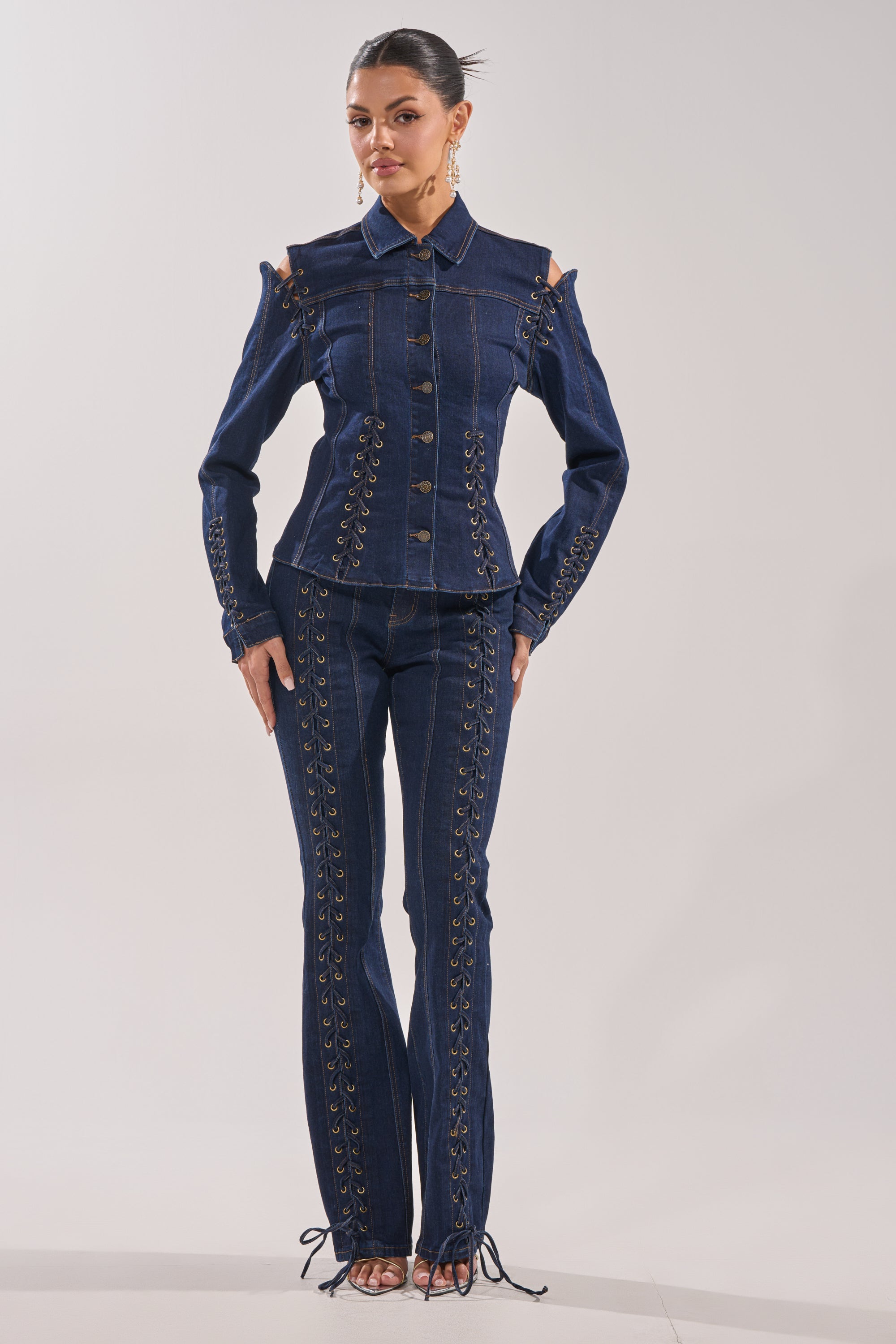A woman poses confidently in the THATS ON ME FLARE DENIM PANT, styled with a matching dark denim jacket featuring lace-up details and cut-out shoulders against a plain light background. 1