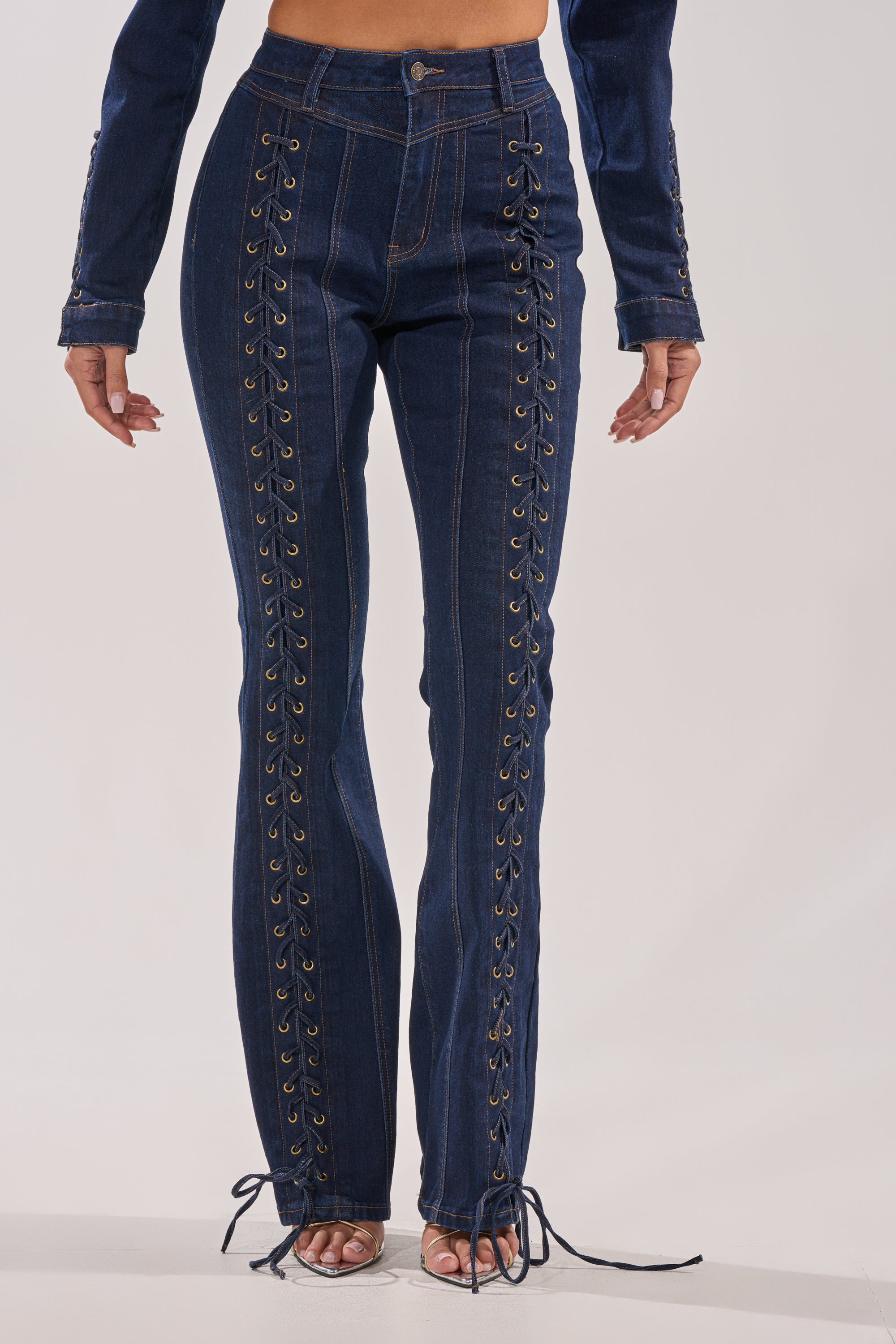 Wearing THATS ON ME FLARE DENIM PANT in dark blue, featuring a high-rise fit, lace-up details on front and sides, fitted silhouette, flared legs, and exposed ankle ties; only the lower torso and legs are shown. 2