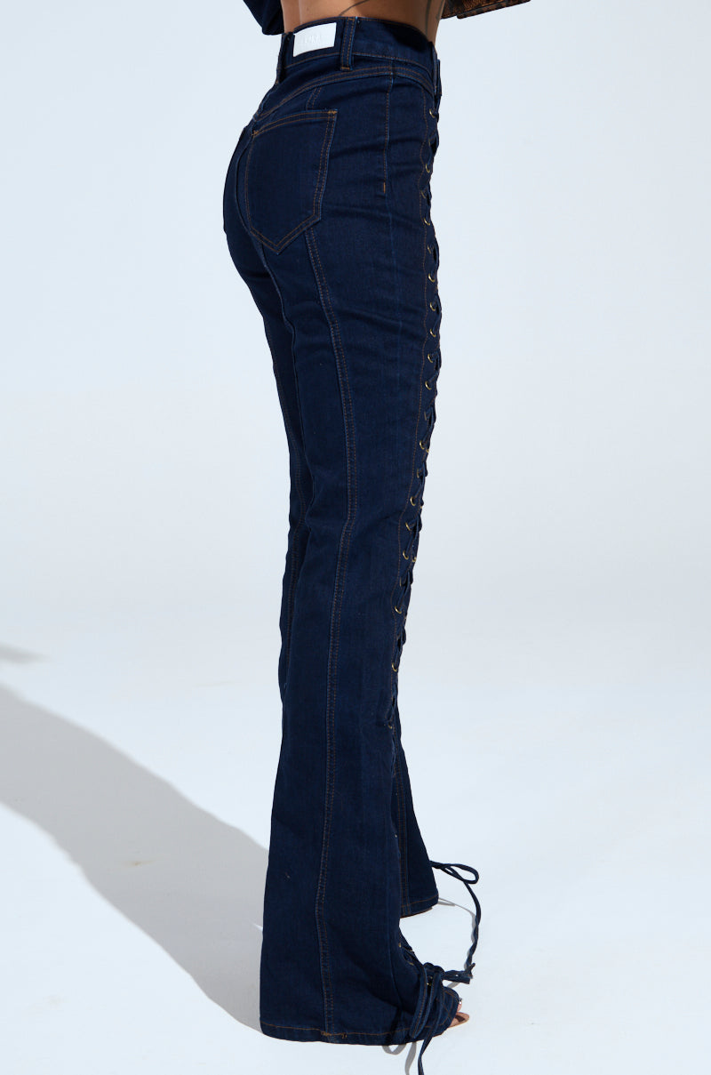 The lower torso of a person, wearing the THATS ON ME FLARE DENIM PANT in dark blue featuring side lacing and visible stitching, stands against a light background. The high-rise flared jeans are paired with a cropped dark top.